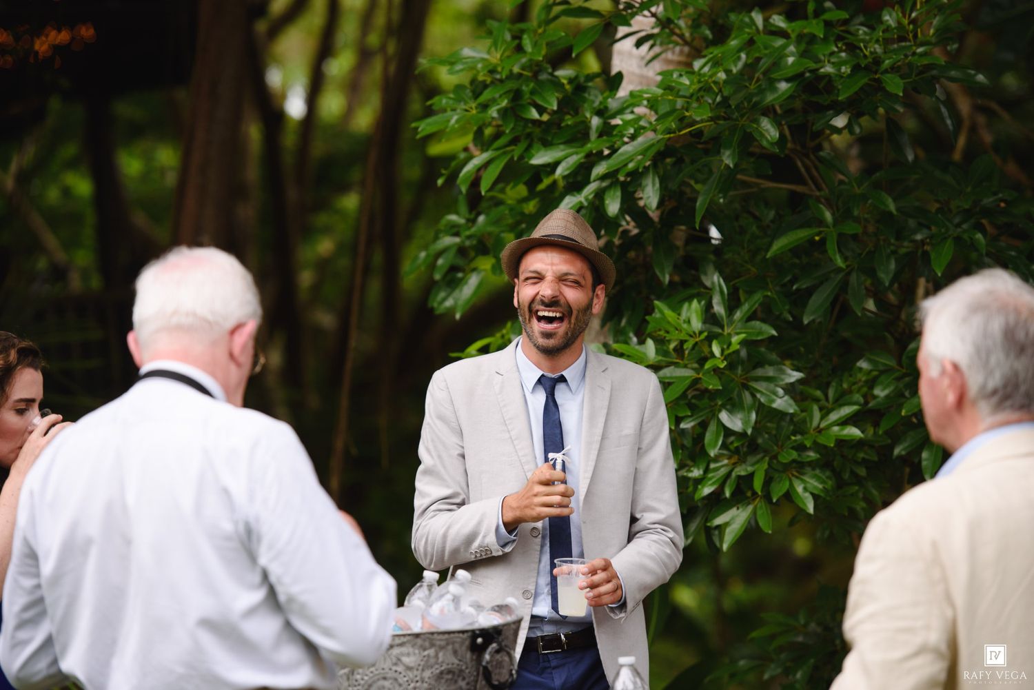 A laughing man in a gray suit and brown hat holds a drink at an outdoor wedding reception surrounded by guests.