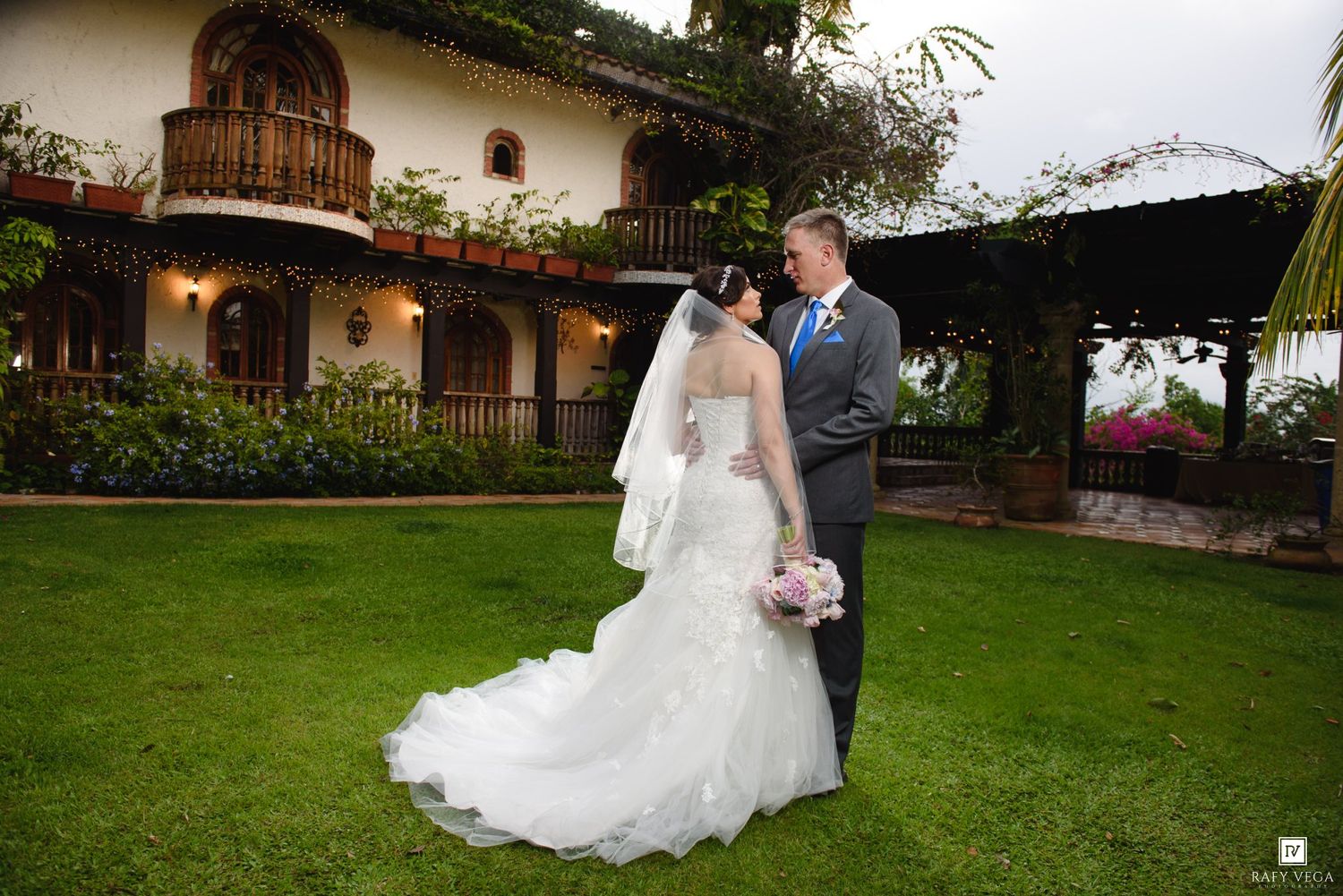 Bride and groom share a romantic moment on lush green lawn outside elegant Spanish-style wedding venue.