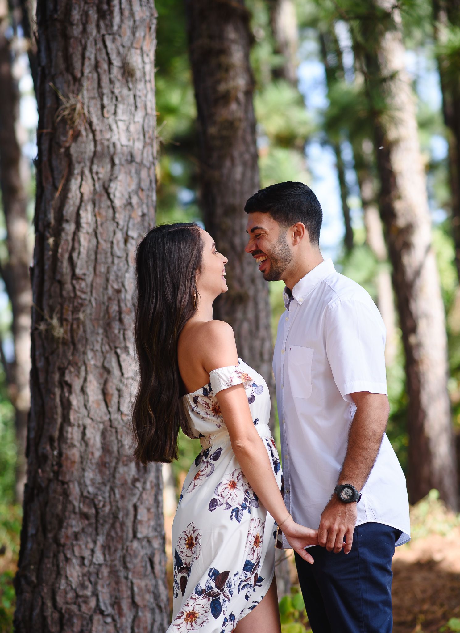 Couple smiling at each other holding hands in a sunlit pine forest, woman in floral dress, man in white shirt.