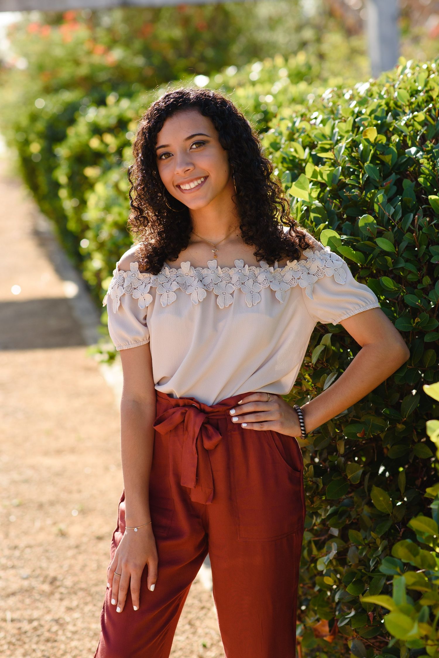 Young woman in off-shoulder floral top and rust-red pants smiles confidently outdoors beside lush green hedges.