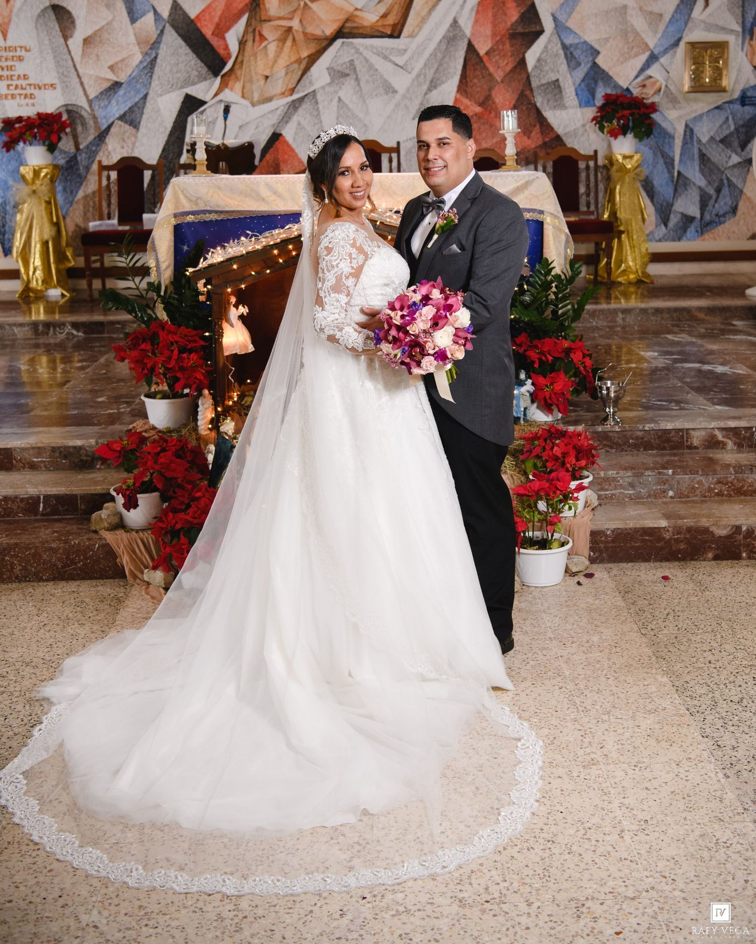 Bride and groom posing at church altar with Christmas poinsettias, bride in lace gown with veil holding pink bouquet.