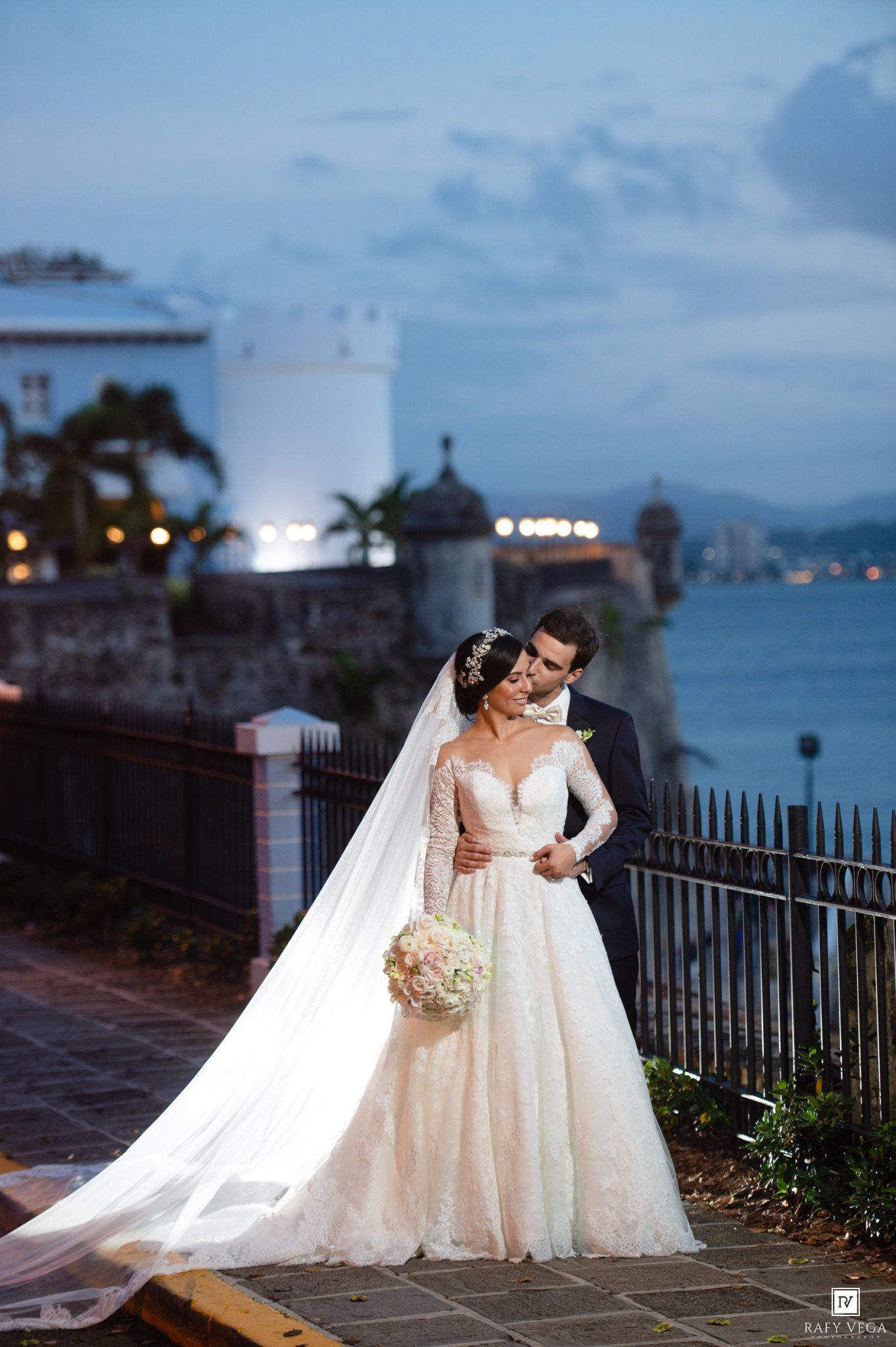 Bride and groom share romantic kiss at dusk by waterfront fortress in elegant lace wedding gown with long veil.