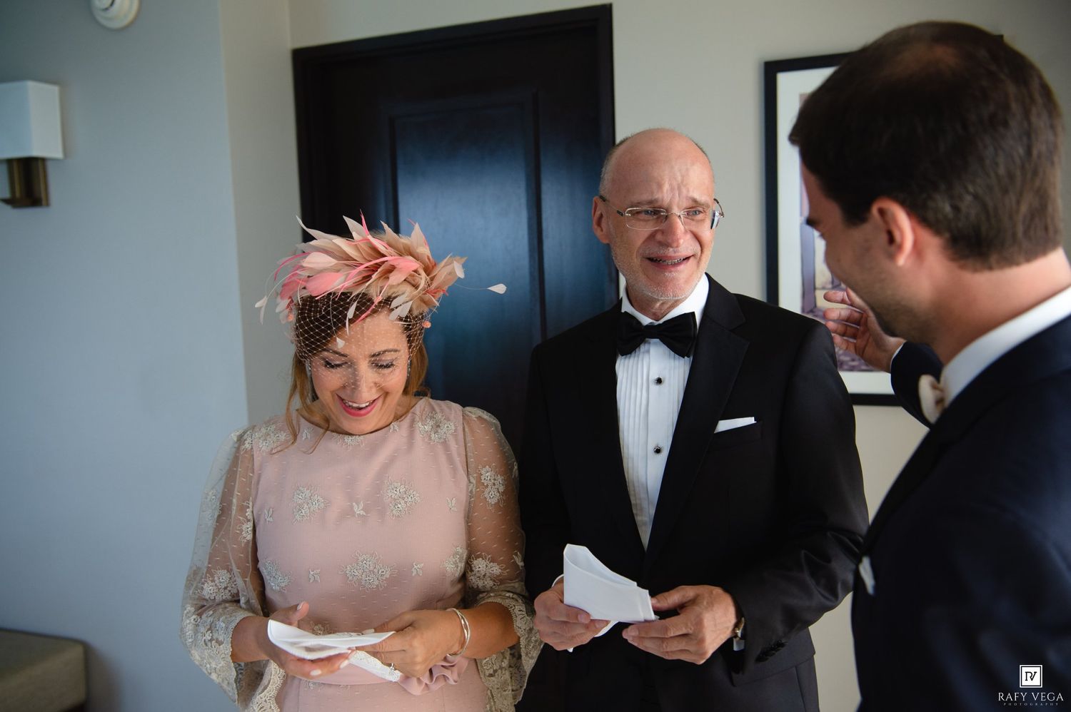 Elegantly dressed couple and man in tuxedo sharing a joyful moment at a formal wedding reception indoors.