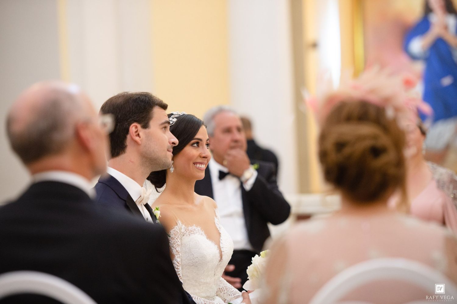 Bride and groom smiling during elegant indoor wedding ceremony surrounded by guests in formal attire.