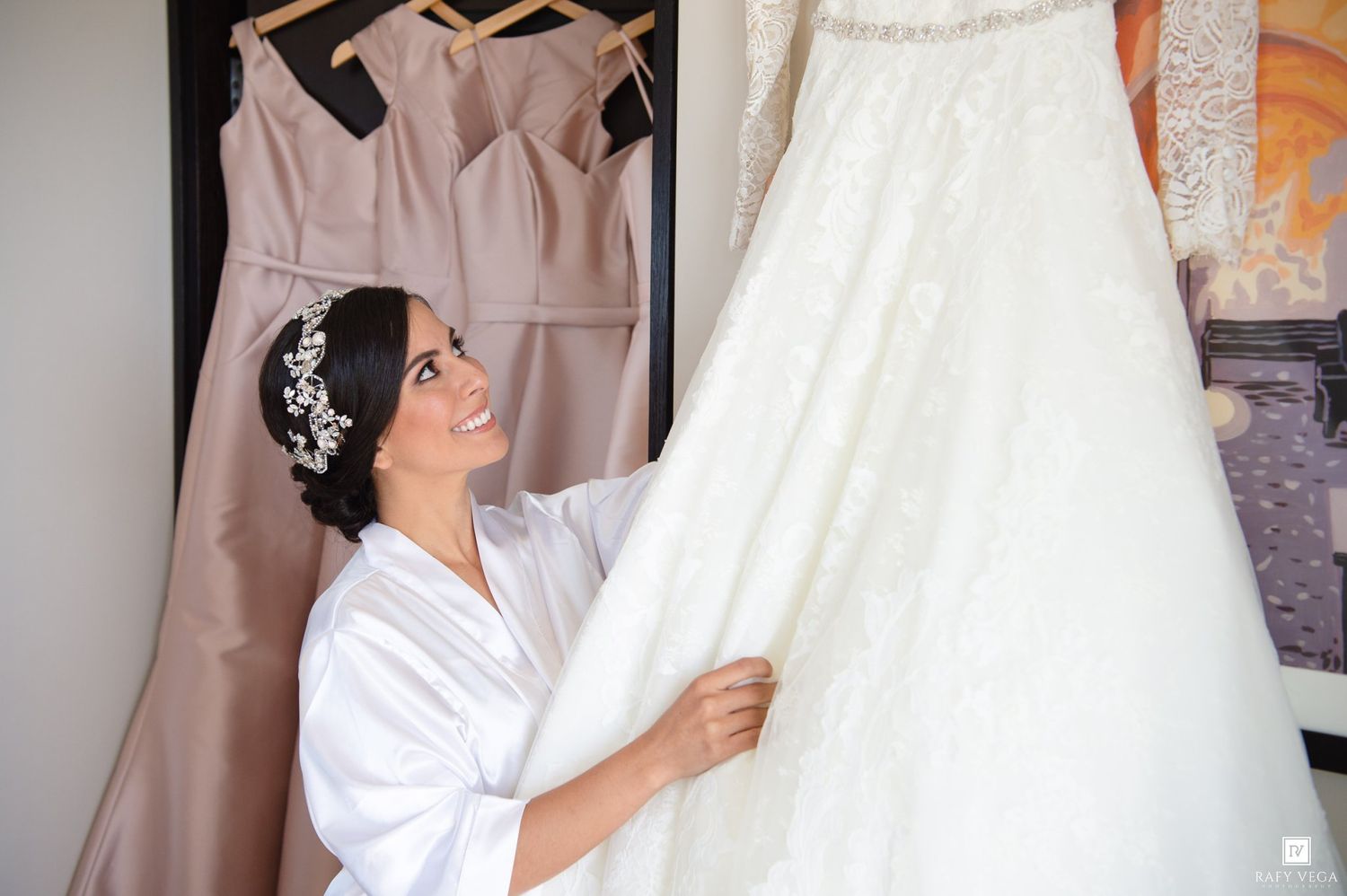 Smiling bride in white robe admiring her lace wedding gown hanging beside blush bridesmaid dresses.