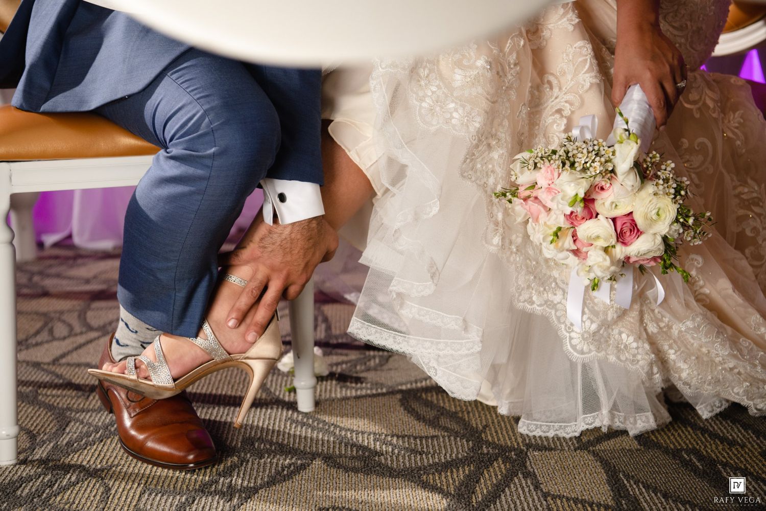 Groom adjusting bride's heels while she holds a pink and white floral bouquet in a wedding dress.