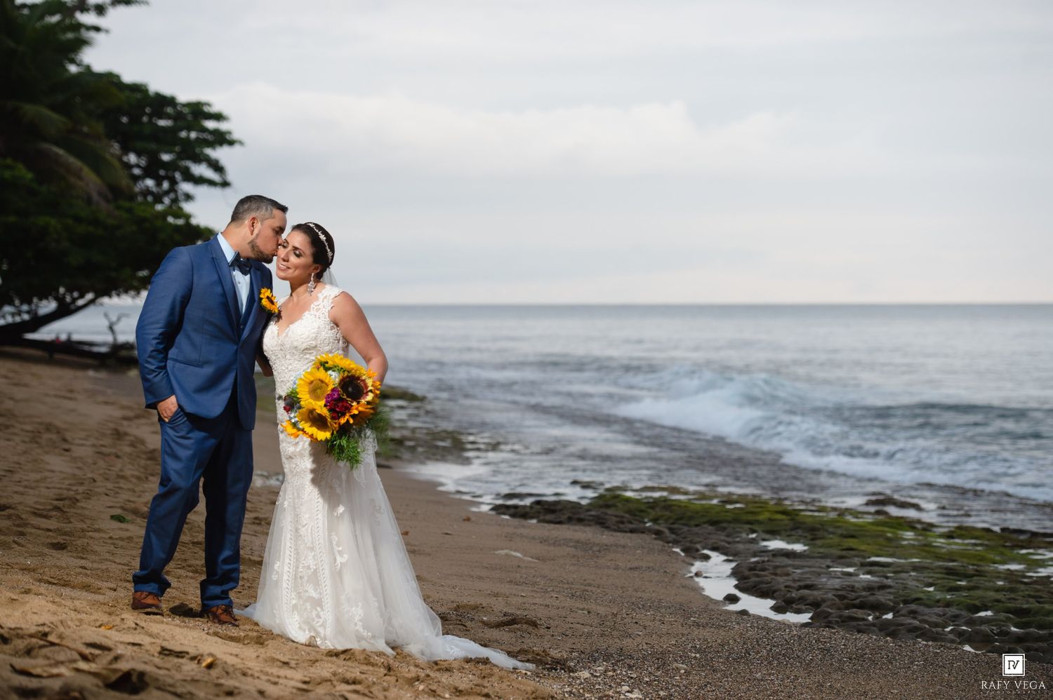 Bride and groom share a romantic kiss on a tropical beach, bride holding vibrant sunflower bouquet in white gown.