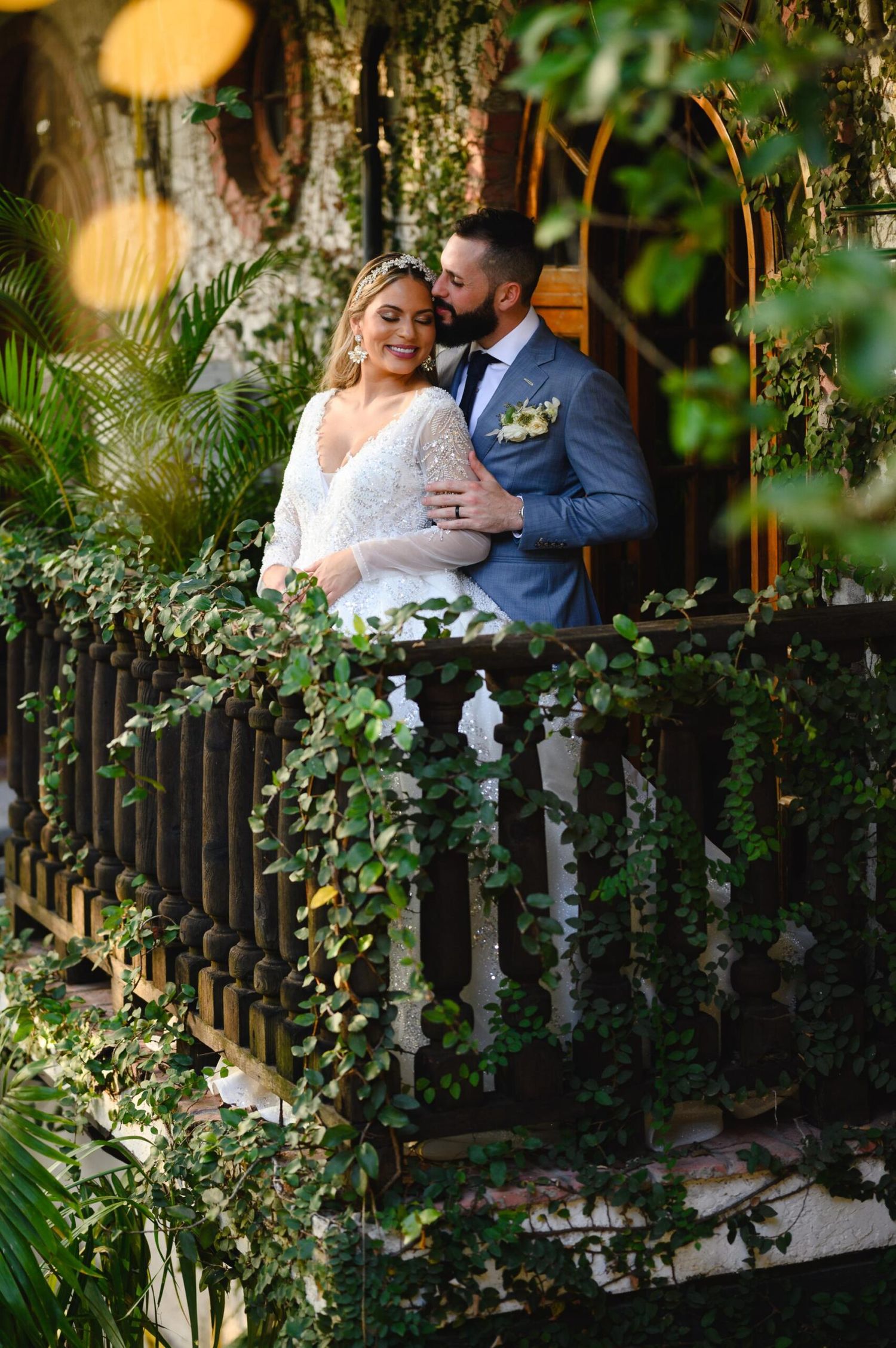 Bride and groom share a romantic moment on an ivy-covered balcony surrounded by lush tropical greenery.