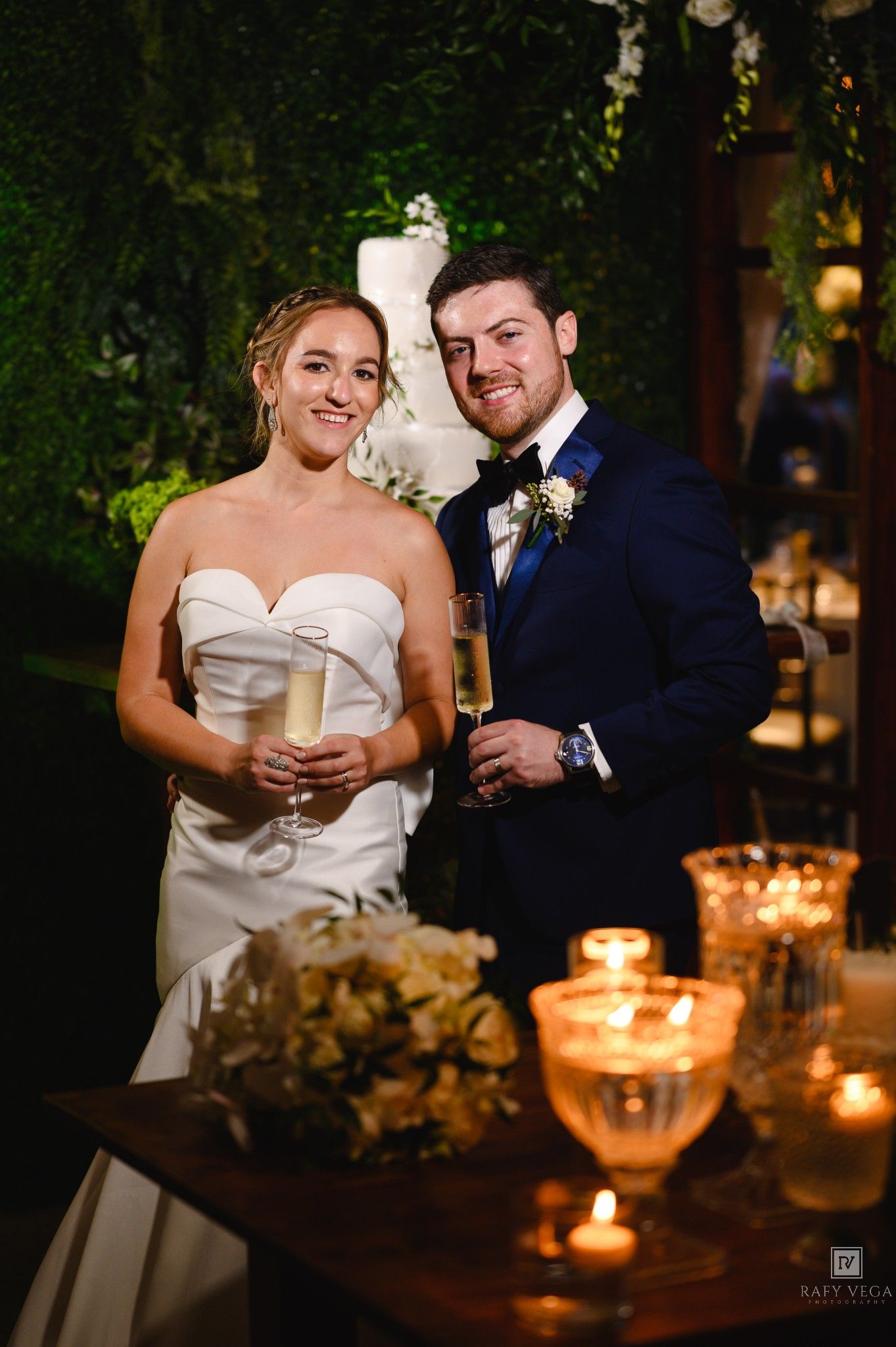 Bride and groom toast with champagne at candlelit sweetheart table, white wedding cake and greenery wall backdrop.