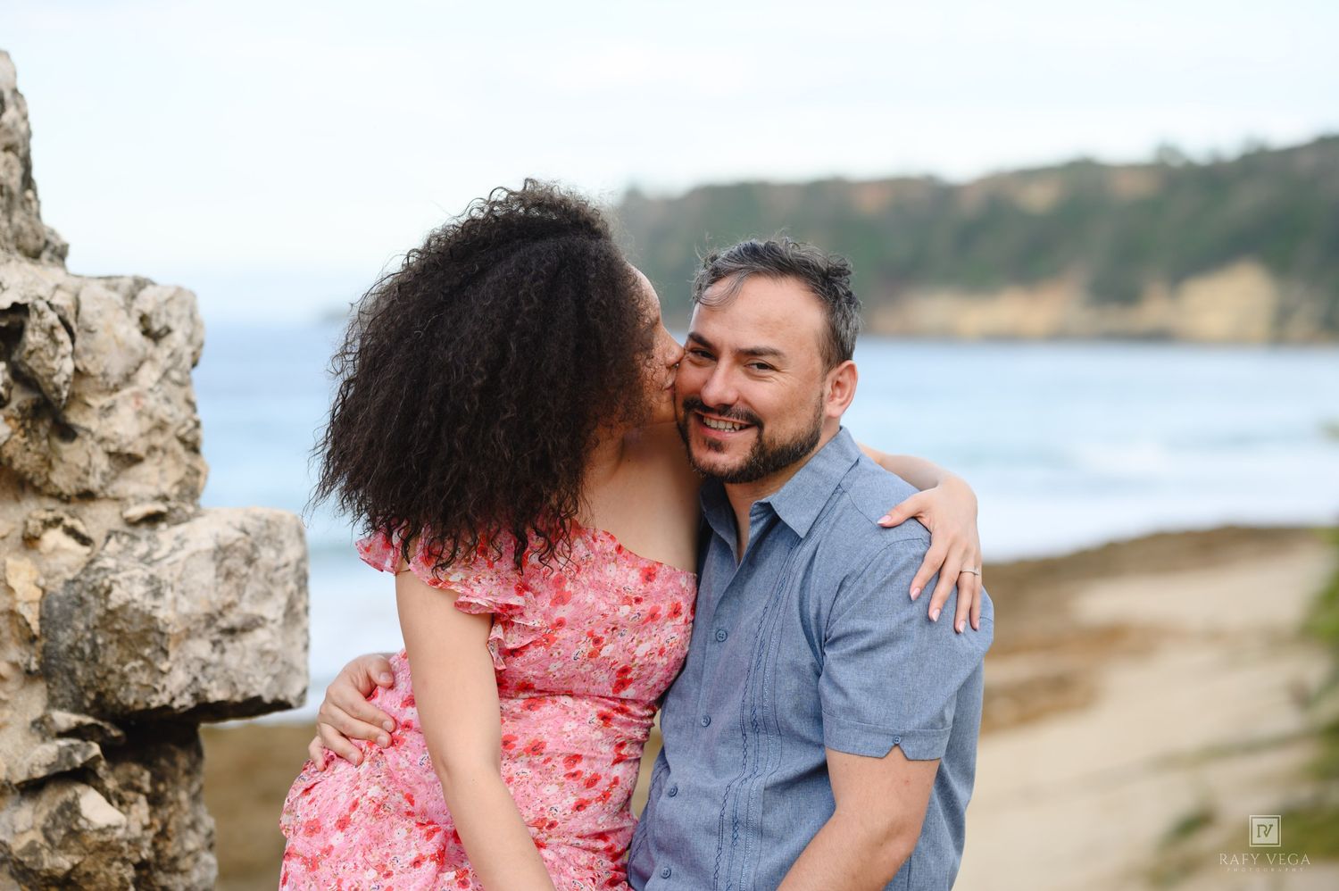 Couple sharing a romantic moment on a scenic beach, woman kissing man's cheek near rustic stone wall.