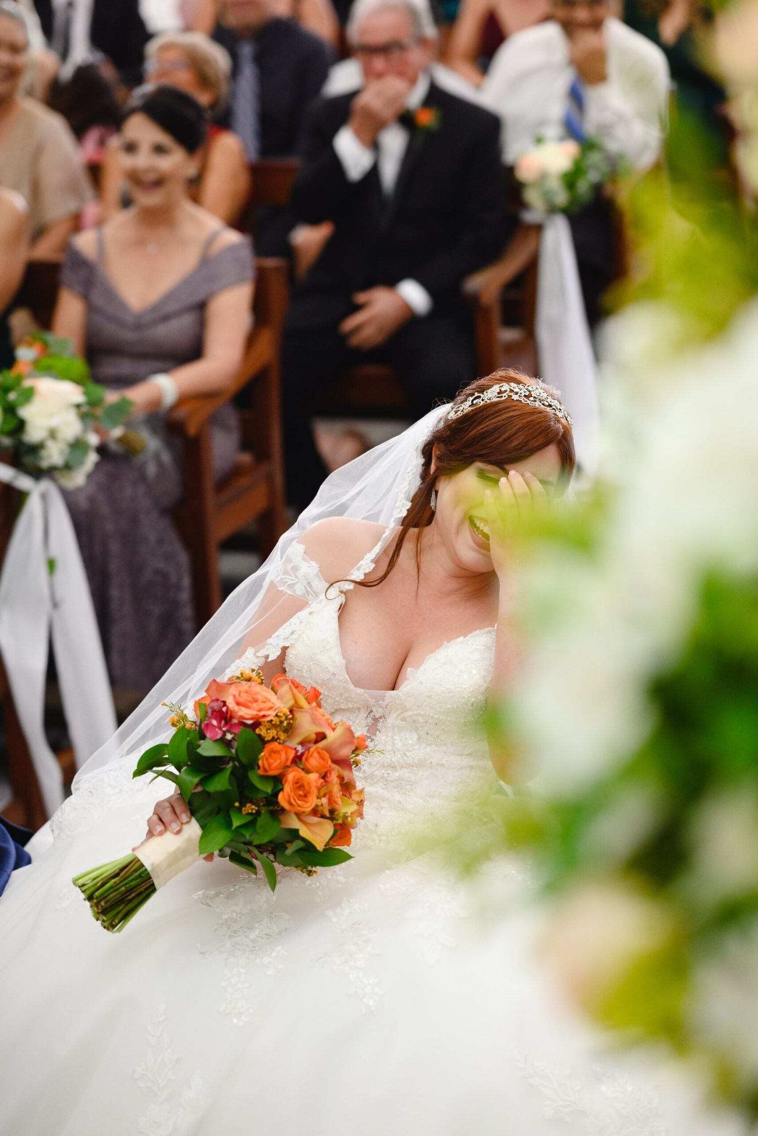 Bride in white lace gown and veil holds orange rose bouquet walking down aisle at wedding ceremony.