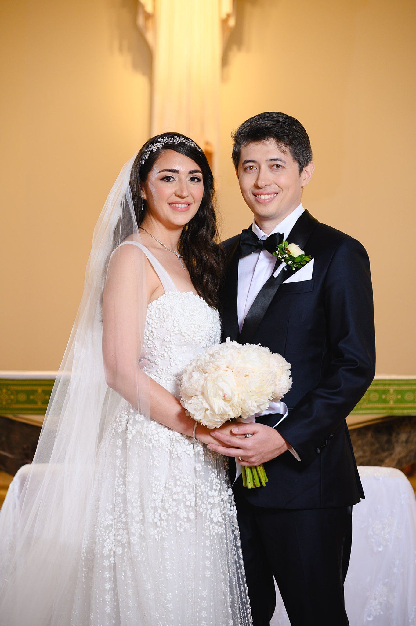 Smiling bride and groom pose together holding a white peony bouquet during their elegant wedding ceremony.