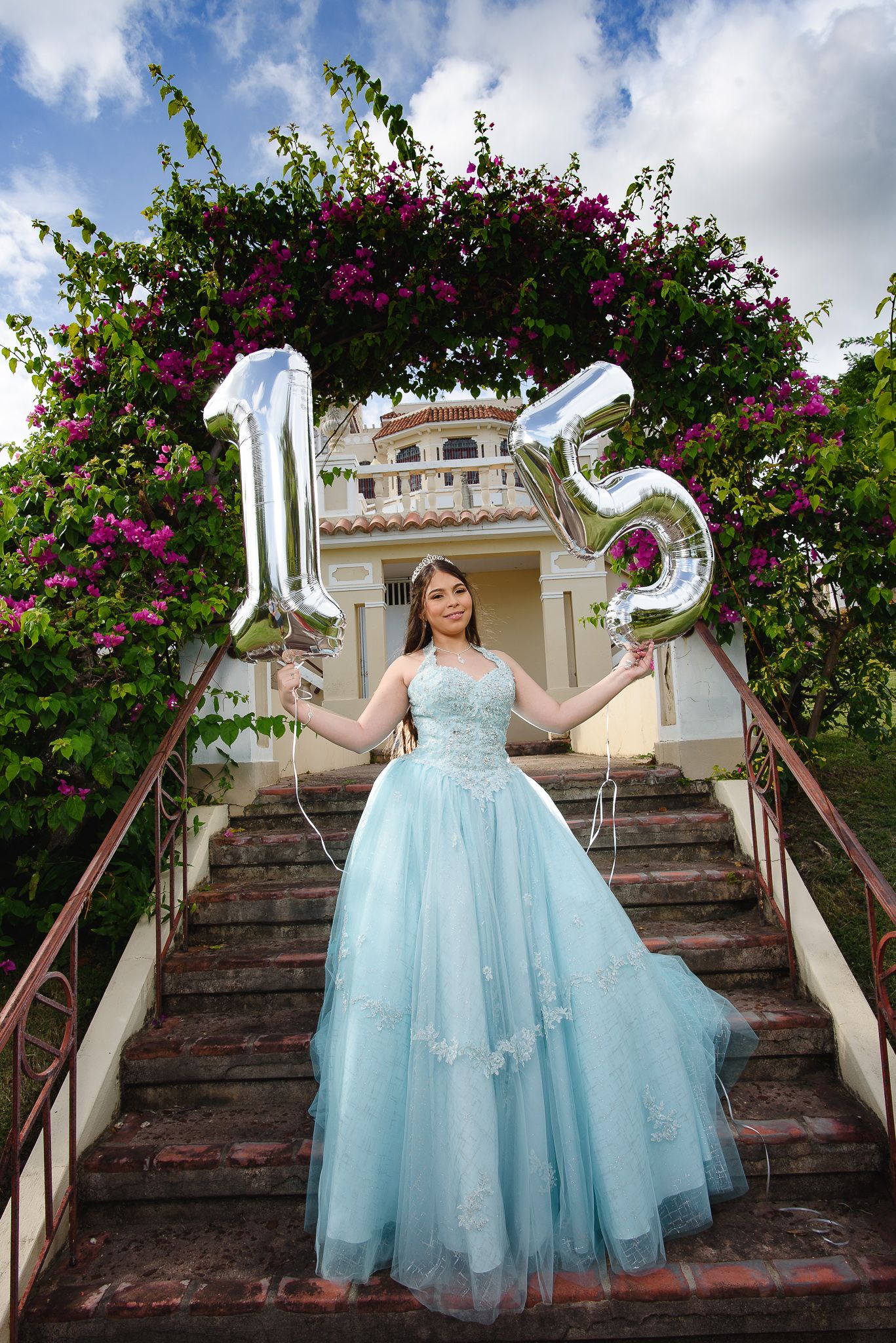 Teen girl in blue quinceañera gown holds silver 15 balloons under floral arch on brick steps.