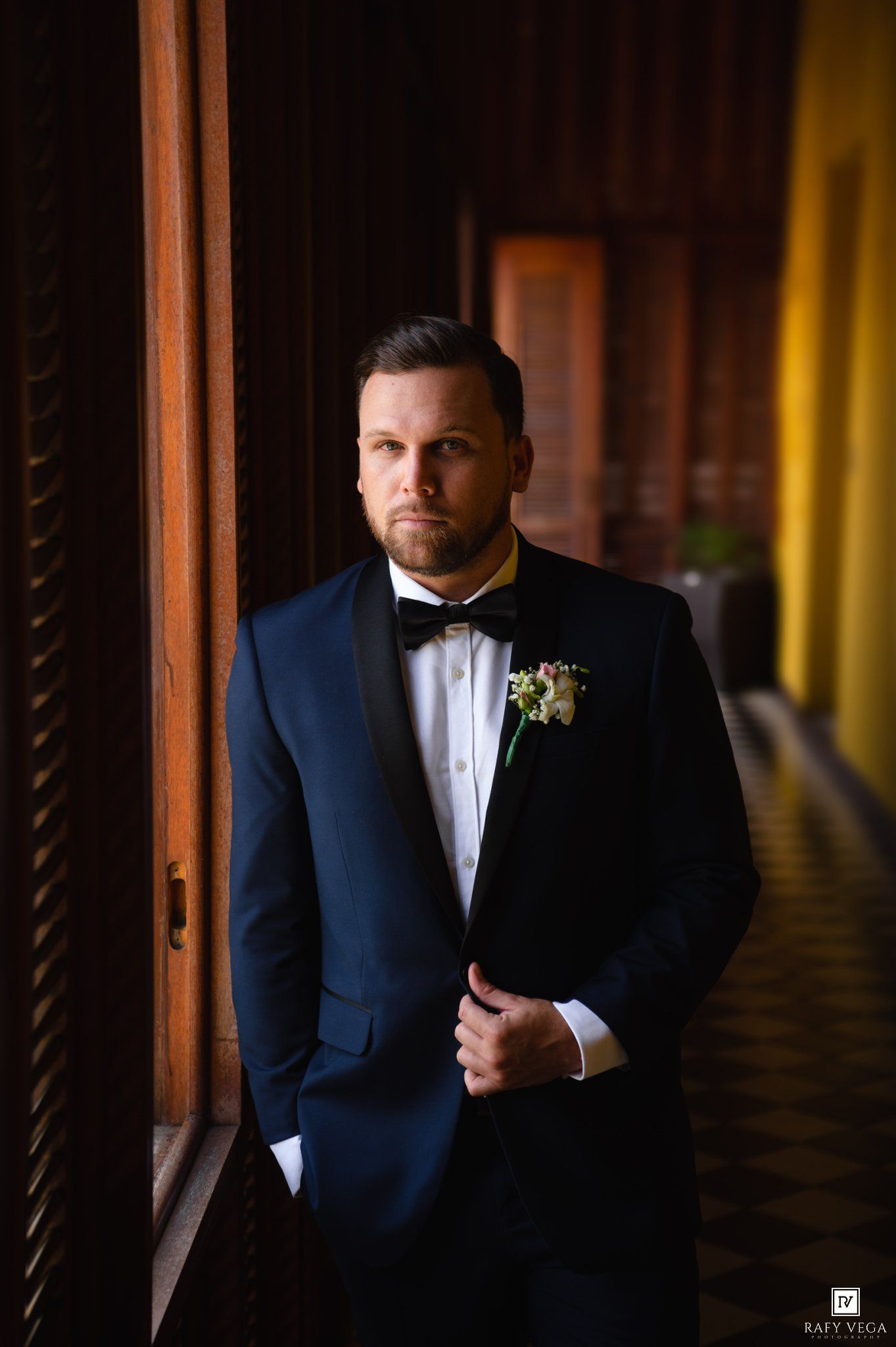 Groom in navy tuxedo with bow tie and boutonniere poses confidently in elegant corridor for wedding portrait.