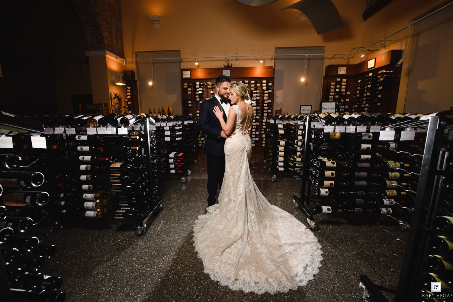 Bride and groom embrace romantically in an elegant wine cellar surrounded by floor-to-ceiling wine racks on their wedding day.