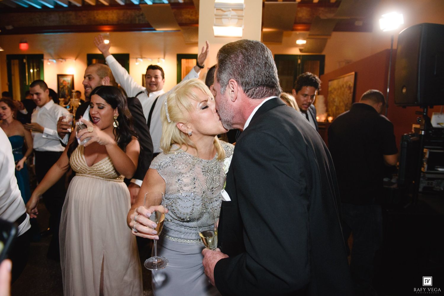Couple sharing a romantic kiss at a wedding reception while guests celebrate on the dance floor behind them.