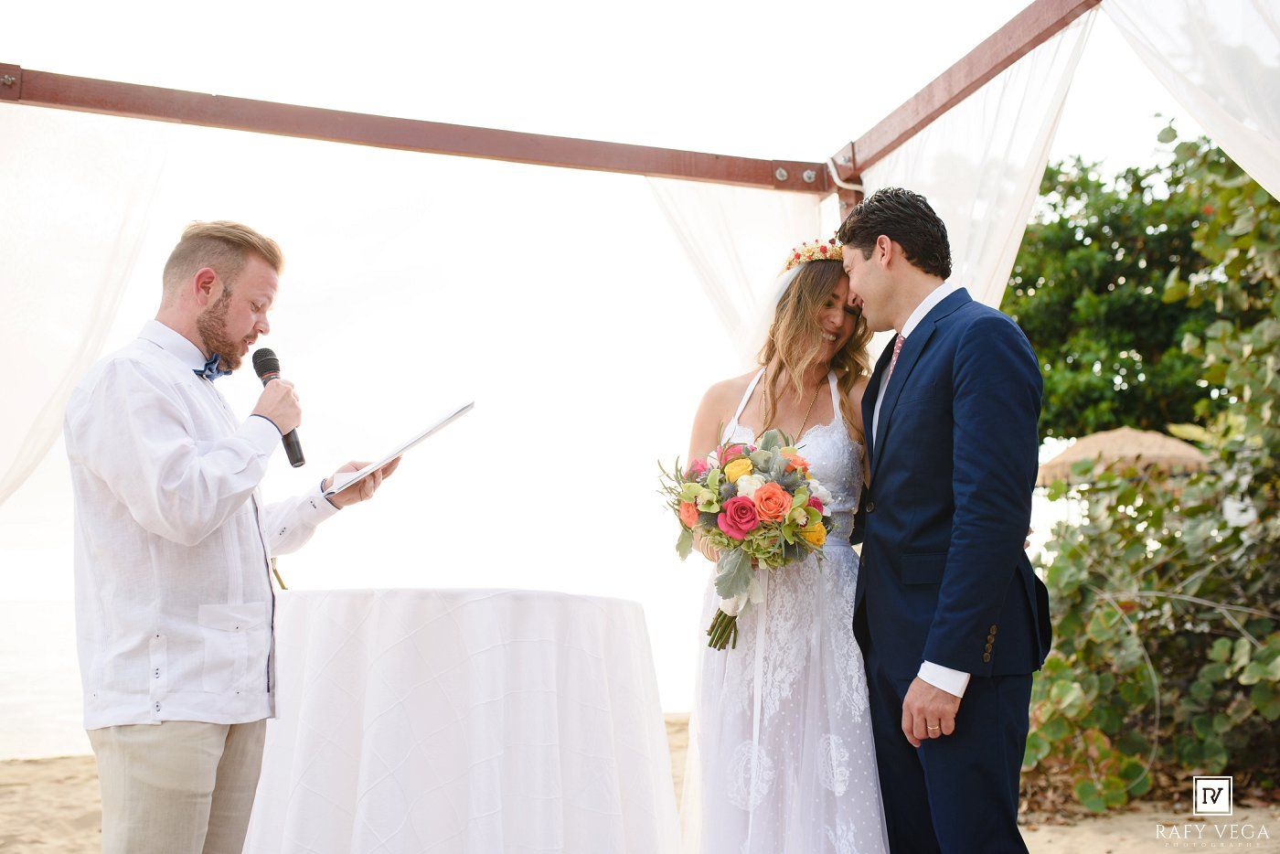 Officiant reads vows to bride and groom during outdoor beach wedding ceremony under white draped arch.
