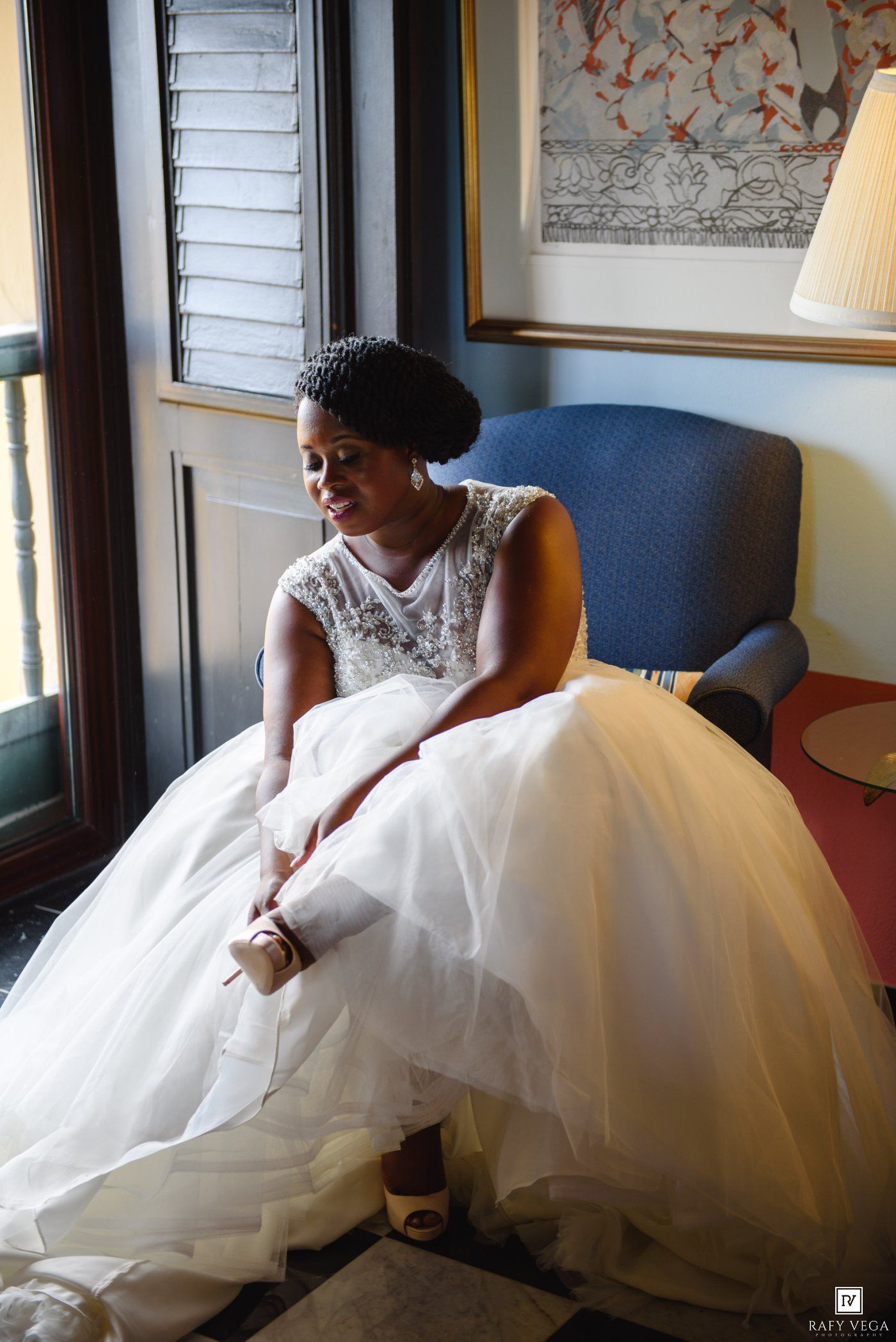 Bride in white ballgown wedding dress sits by window adjusting her shoes, natural light illuminating her elegant look.