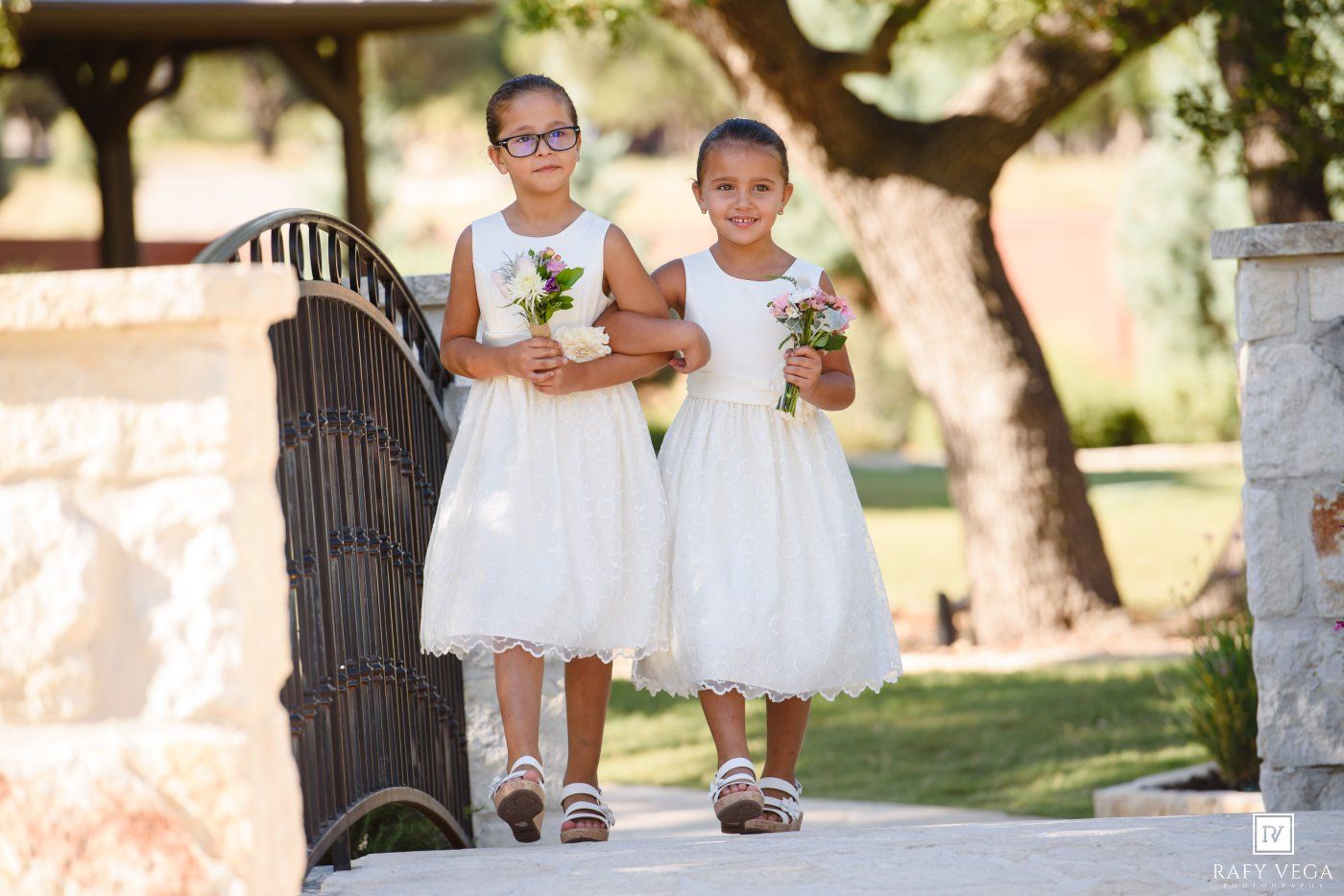 Two young flower girls in white dresses holding bouquets walk along a path at an outdoor wedding ceremony.