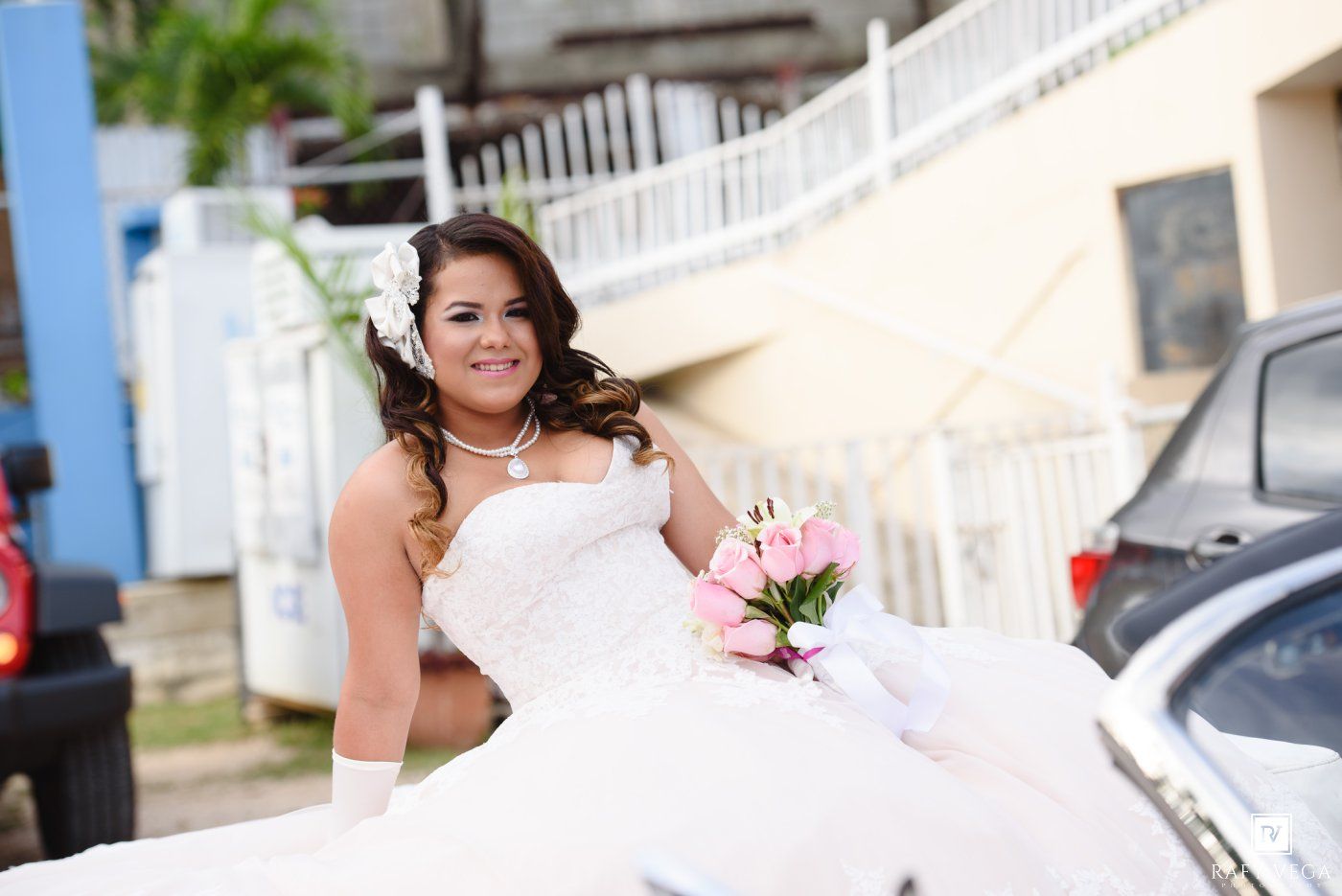 Bride in white strapless wedding gown holding pink bouquet, posing outdoors near a white car on her wedding day.