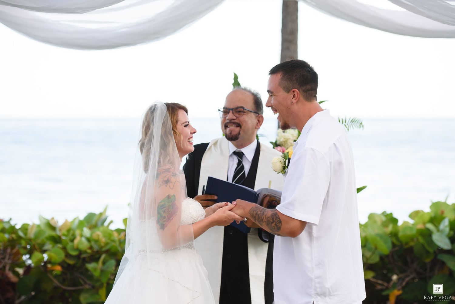 Beach wedding ceremony with bride in white veil, groom in white shirt, and officiant holding book by the ocean.