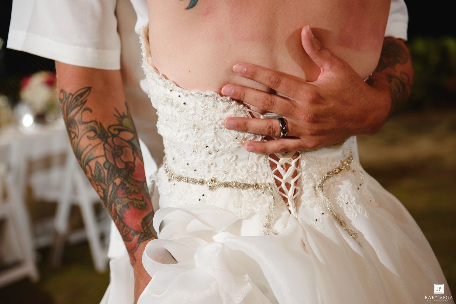 Close-up of bride in beaded wedding dress with groom's tattooed arms embracing her waist at outdoor wedding.