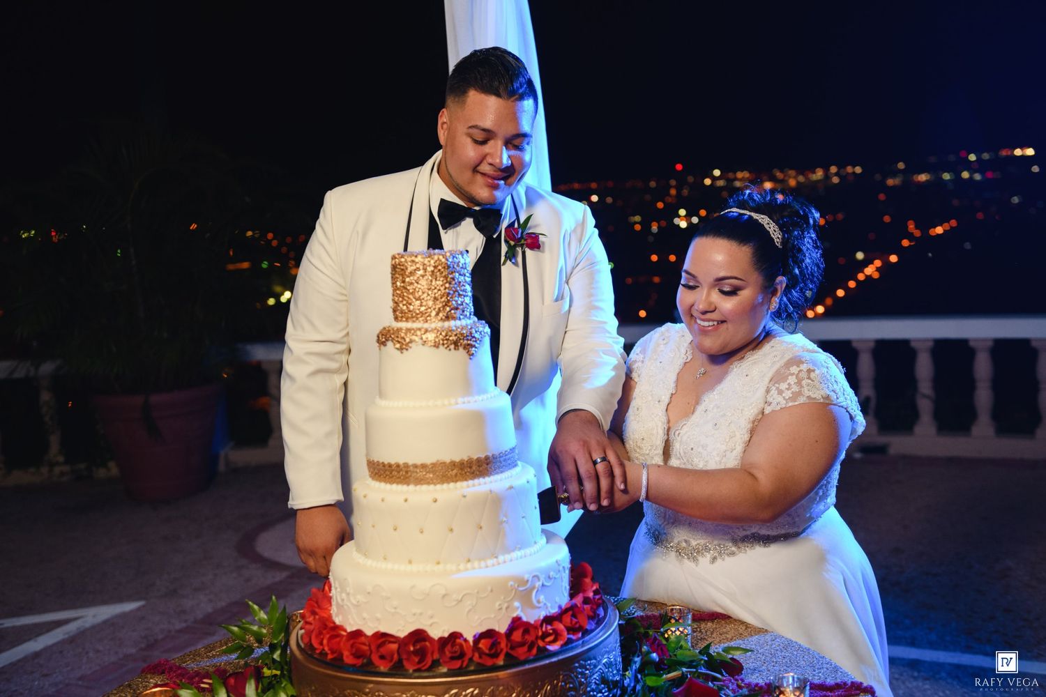 Bride and groom cutting a elegant four-tier white and gold wedding cake outdoors at night with city lights behind.