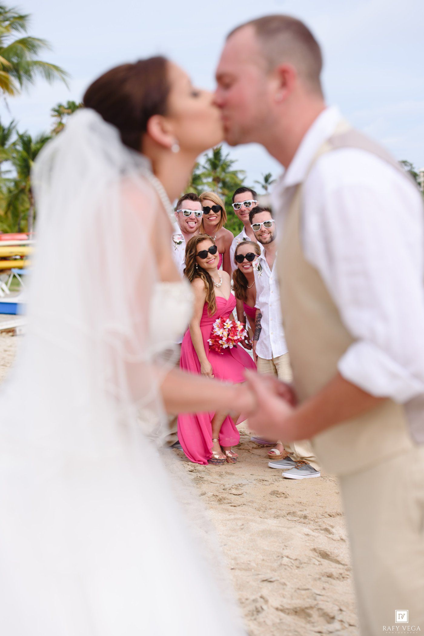 Bride and groom share a beach kiss while wedding party watches and smiles in pink dresses and tan suits.