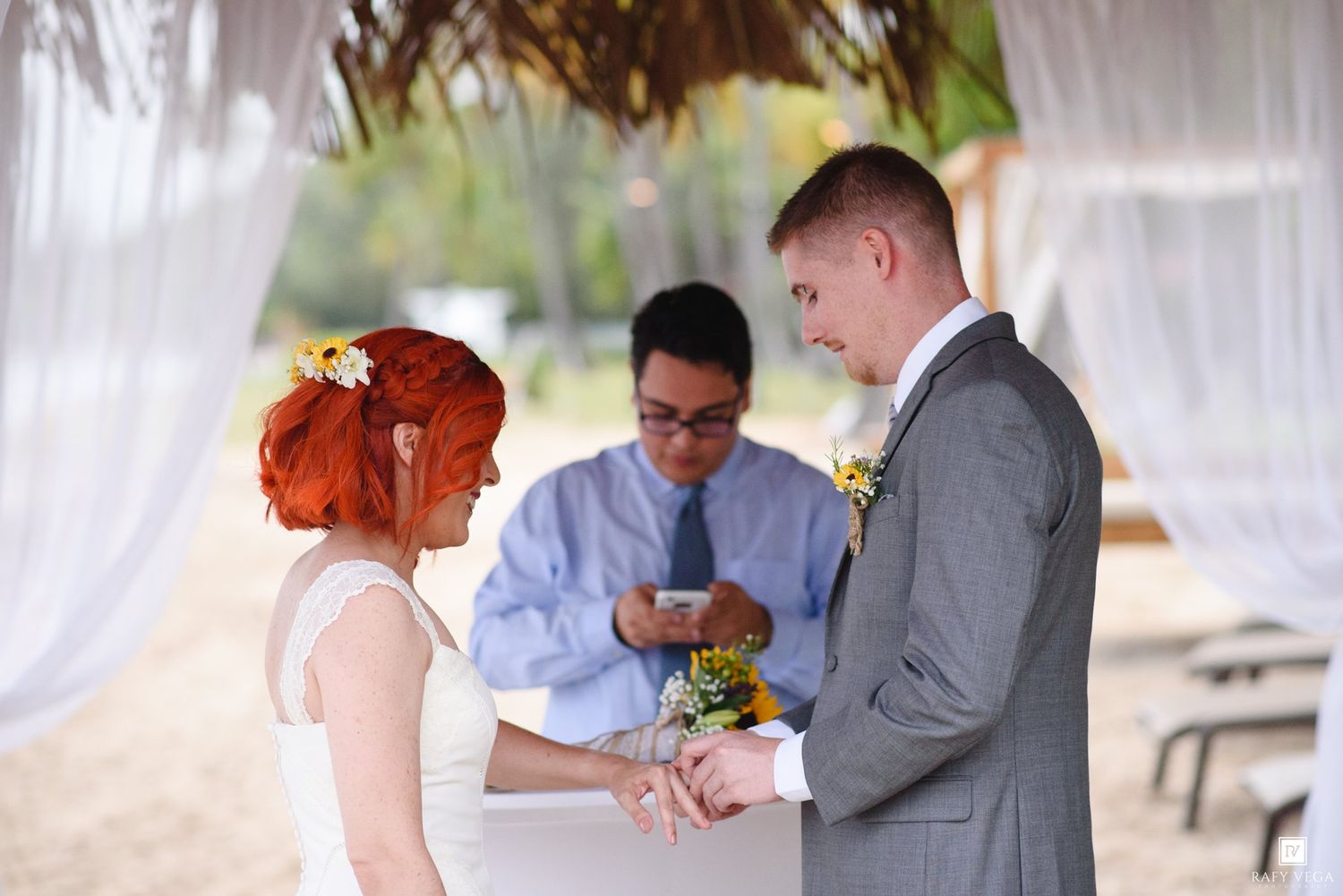 Bride and groom exchange rings during an outdoor beach wedding ceremony with an officiant reading vows.