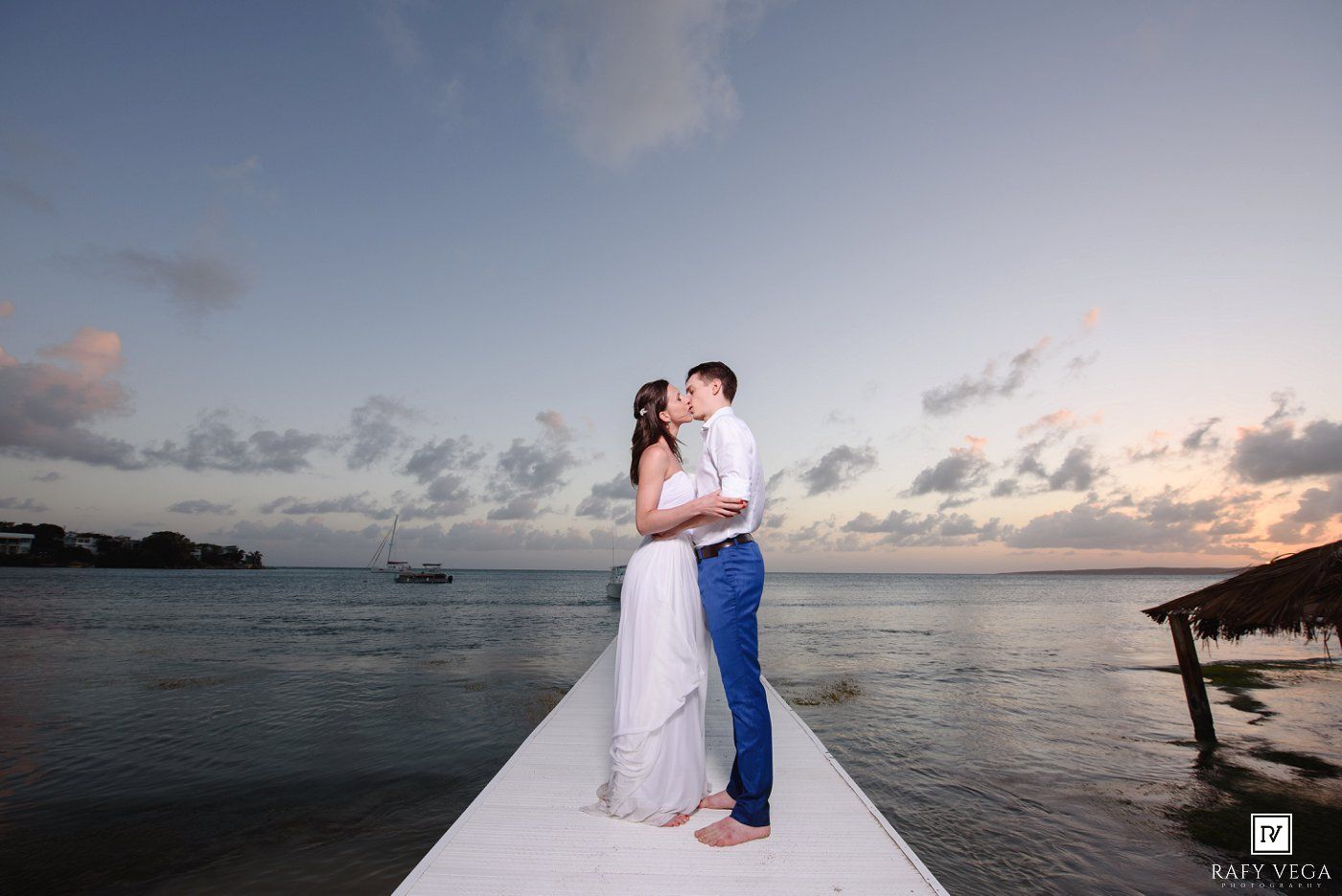 Couple shares romantic embrace on a white dock at sunset over calm tropical waters during their beach wedding.