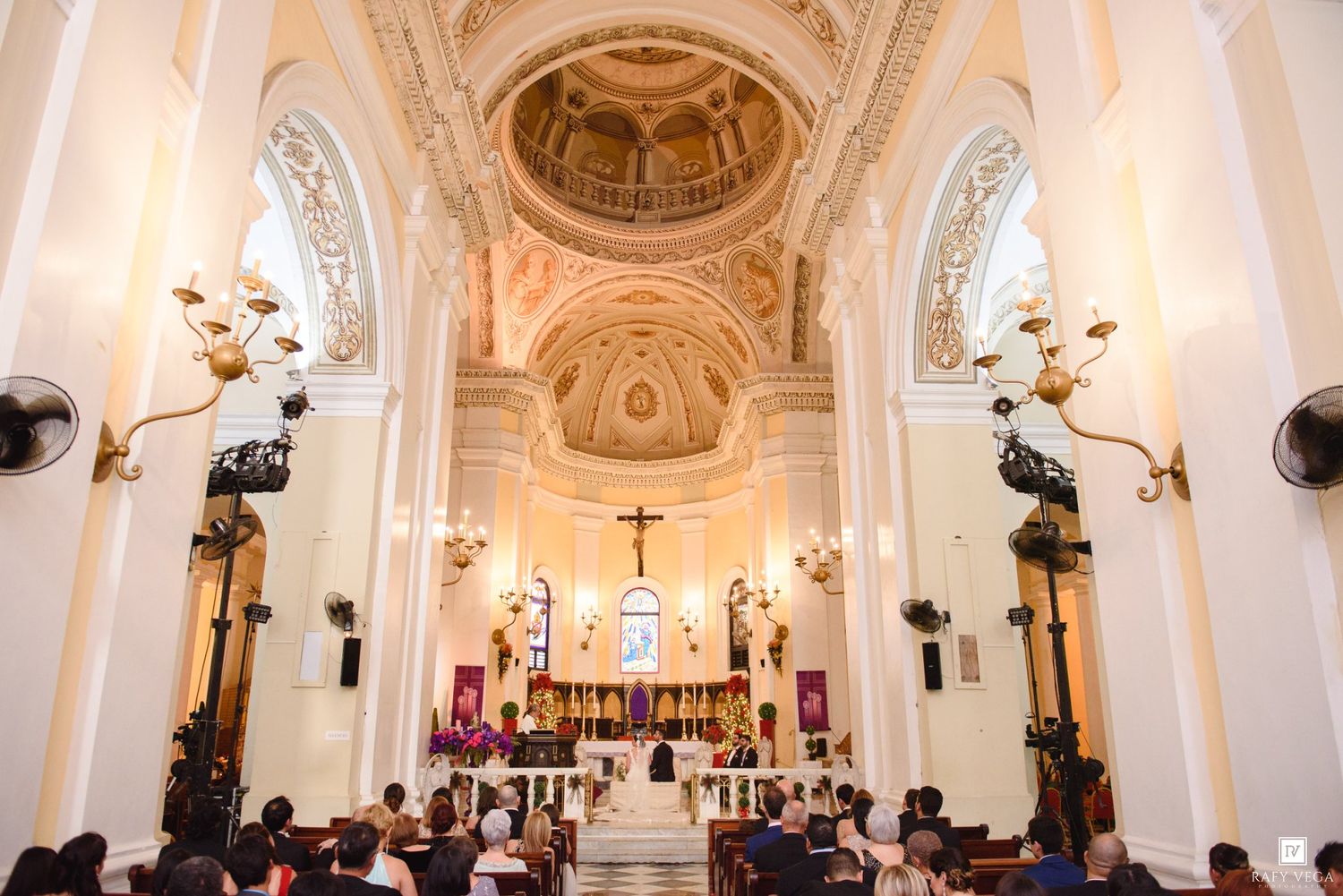 Elegant Catholic church wedding ceremony with ornate gold altar, domed ceiling, and guests seated in wooden pews.