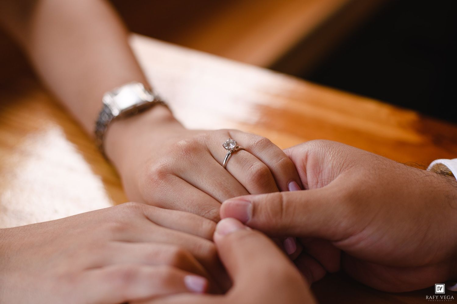 Close-up of hands holding with a diamond engagement ring on finger and silver watch on wrist on wooden table.