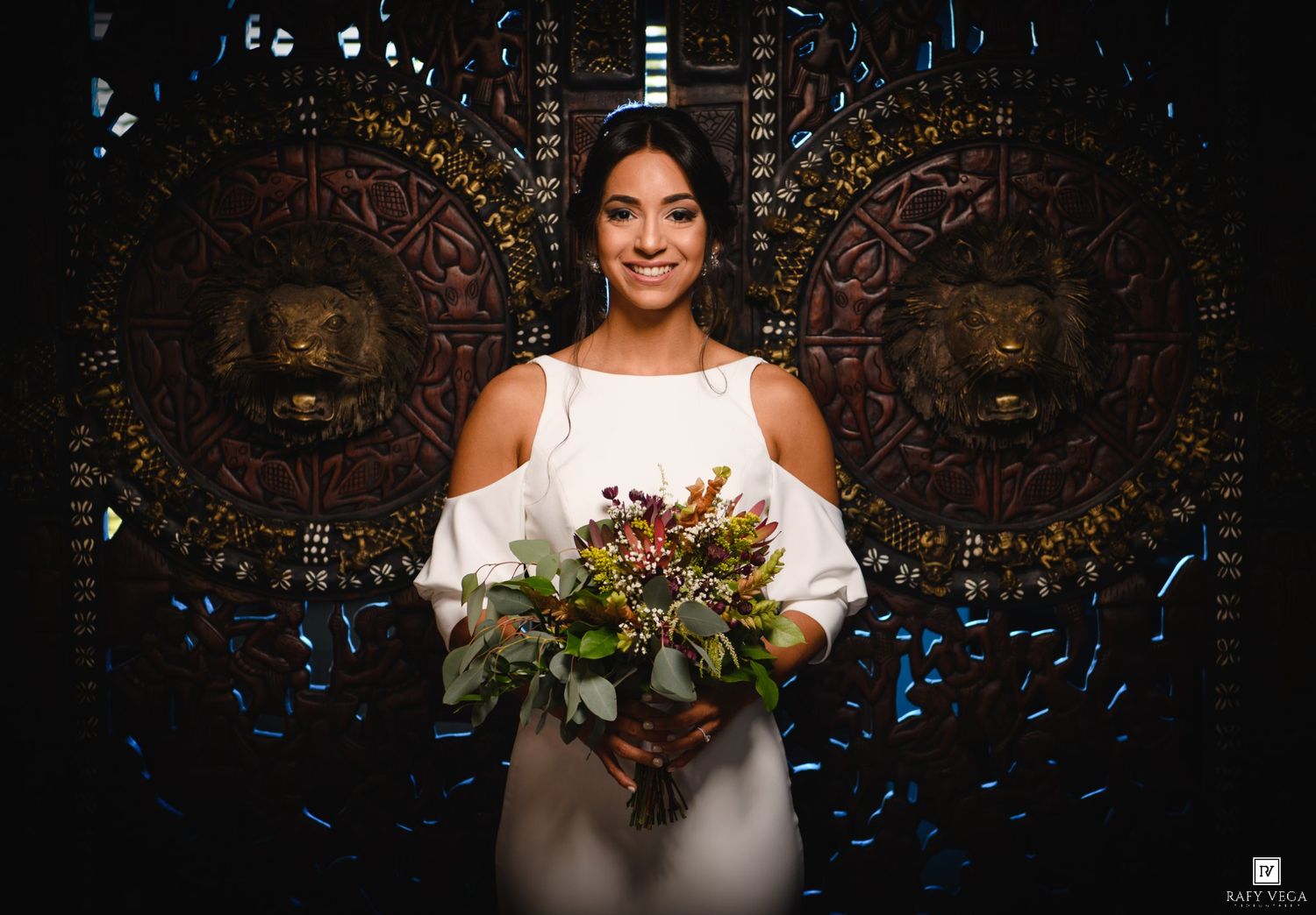 Smiling bride in white dress holding a colorful bouquet in front of ornate carved lion décor.