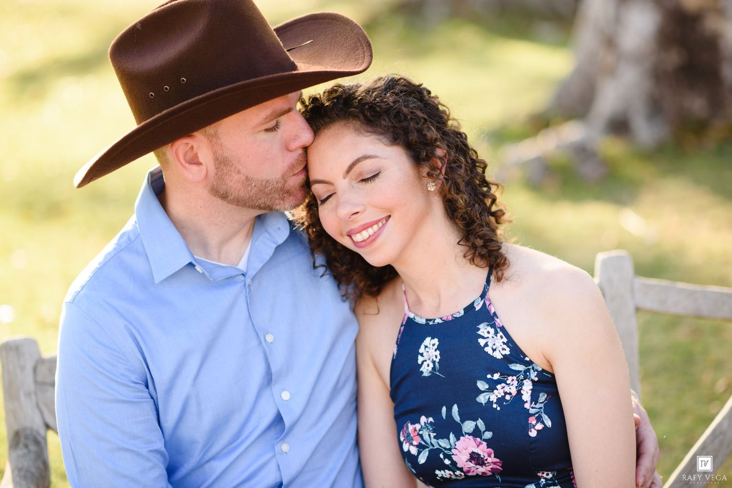 A man in a cowboy hat kisses a smiling woman's cheek outdoors near a wooden fence.