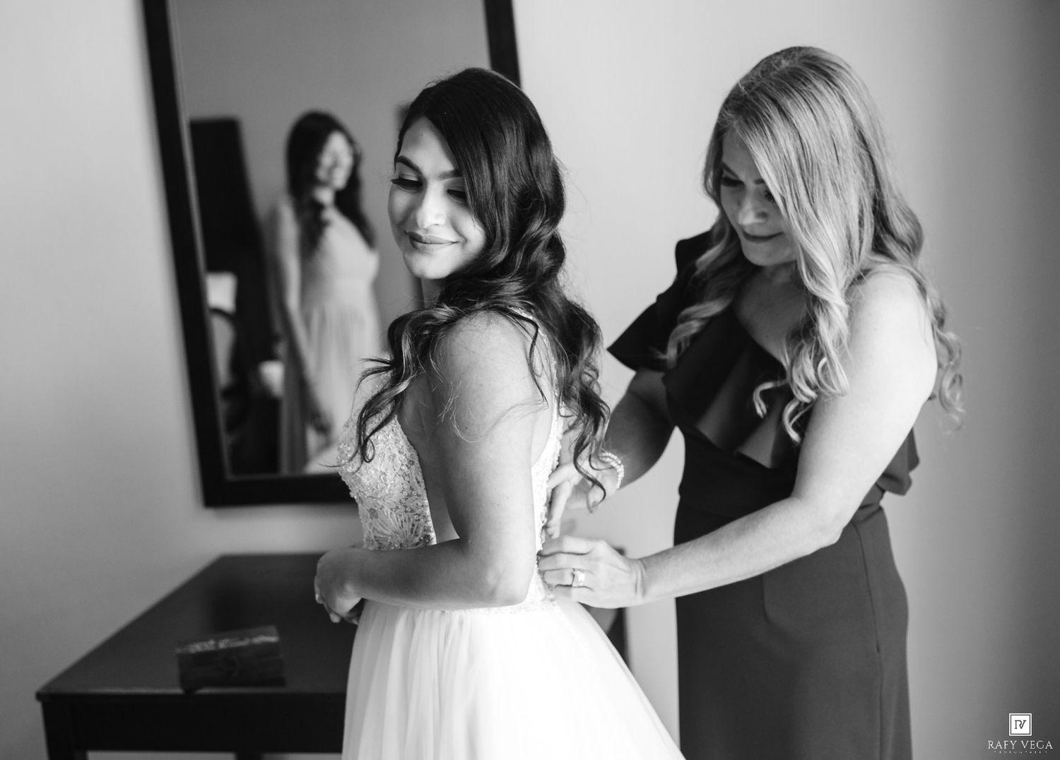 Black and white photo of a bride getting her wedding dress fastened by a bridesmaid, with their reflection visible in a mirror.