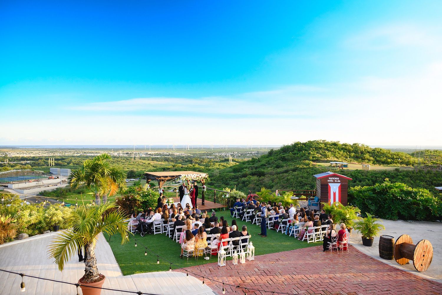 Outdoor wedding ceremony on a hilltop venue with panoramic views, white chairs, and lush greenery under a bright blue sky.