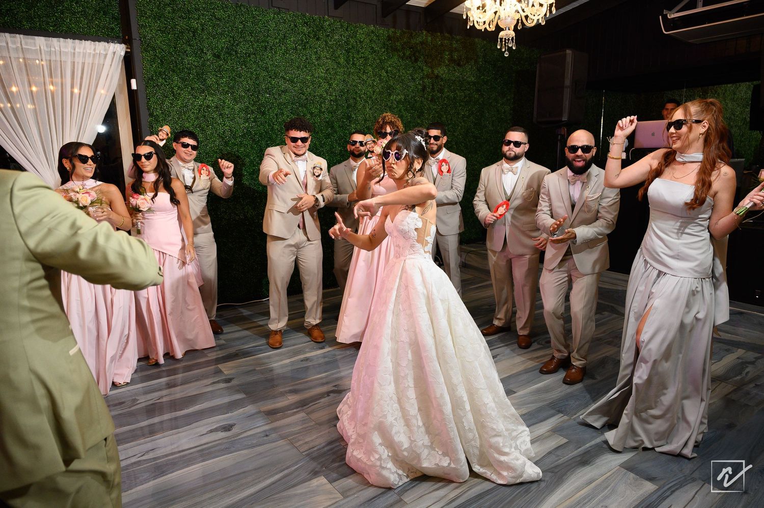 Bride in a white gown dances joyfully on the dance floor as groomsmen in tan suits and bridesmaids in pink dresses cheer around her.