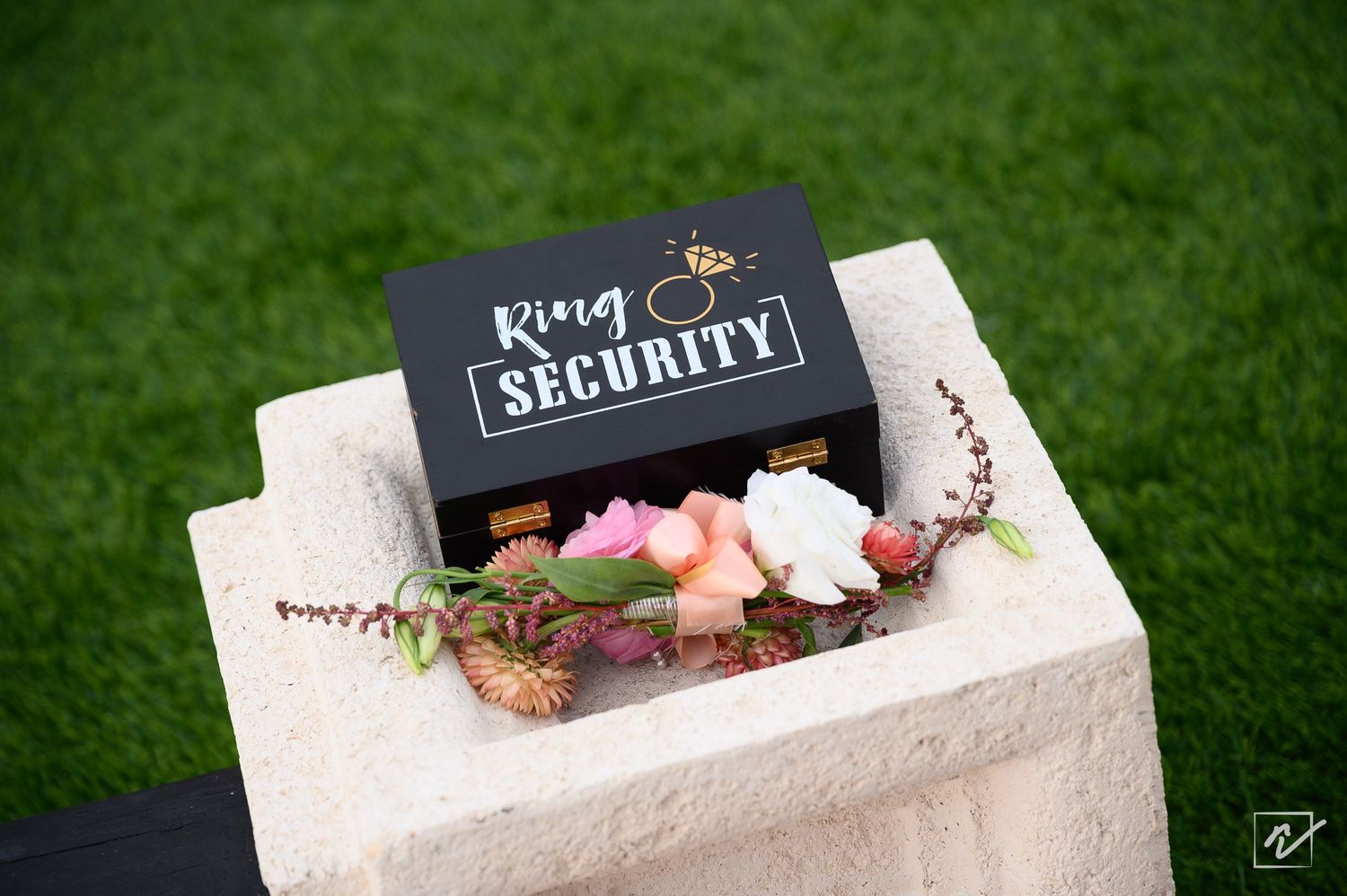 Black wooden ring security box with a gold ring logo, decorated with pink and peach flowers, resting on a stone pedestal over green grass.