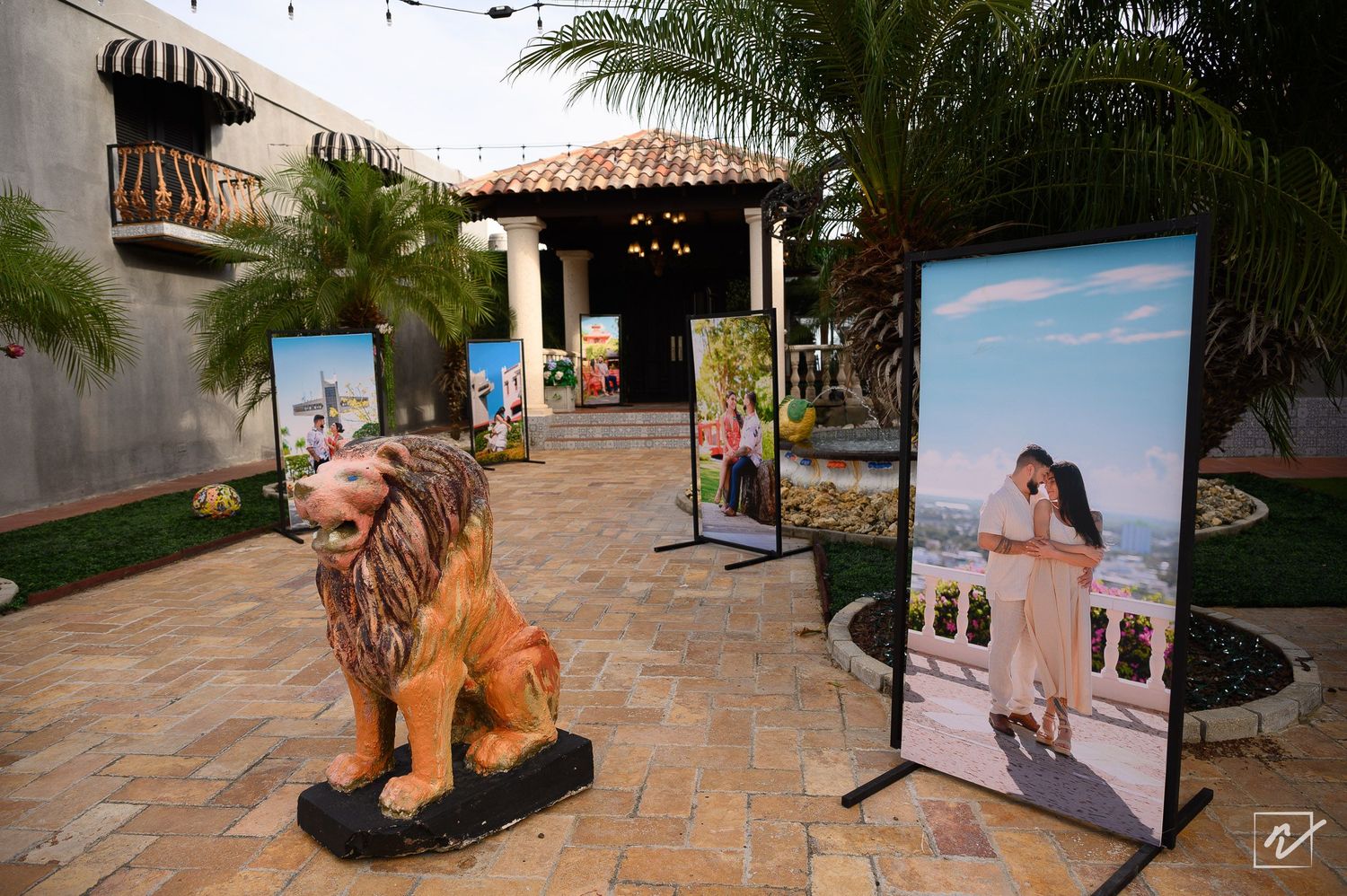 Lion statue and framed couple photo displays on a brick patio with palm trees at an elegant outdoor wedding venue.