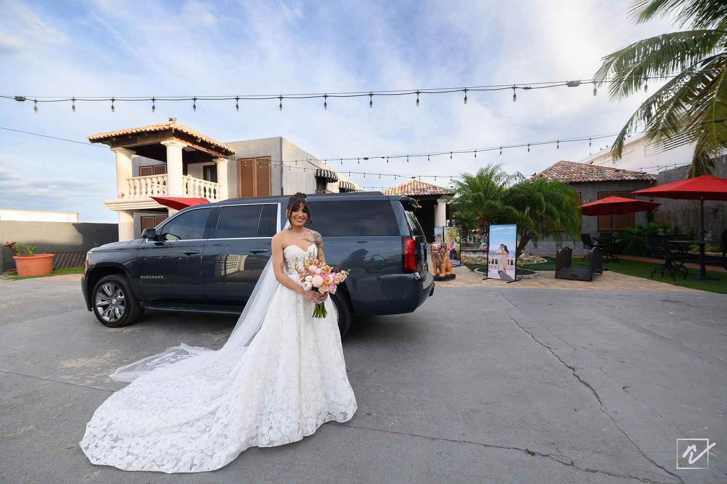 Bride in a long white lace wedding gown holding a bouquet beside a black SUV at an outdoor tropical venue with string lights and palm trees.
