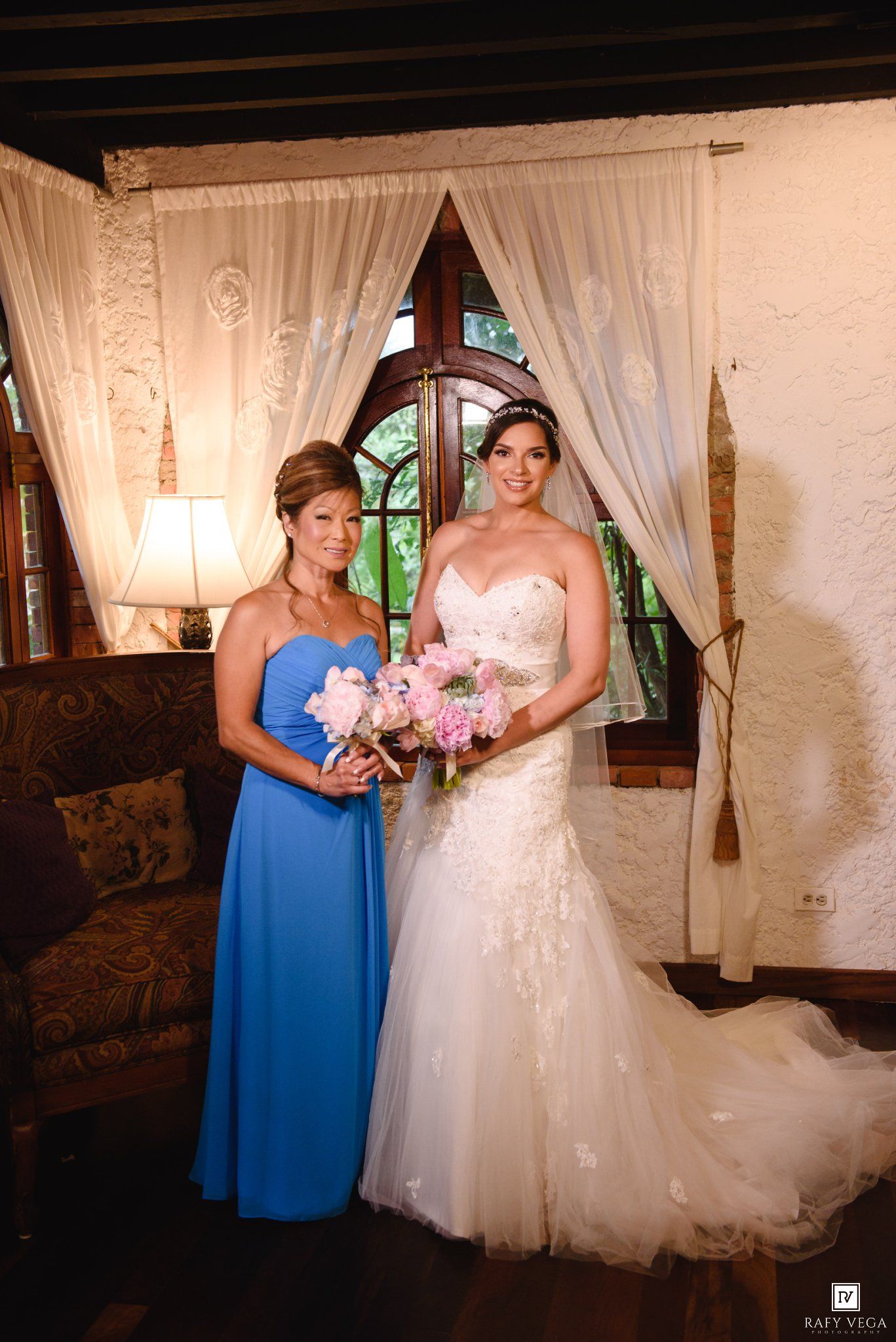 Bride in lace mermaid gown holds pink bouquet posing with bridesmaid in blue dress.