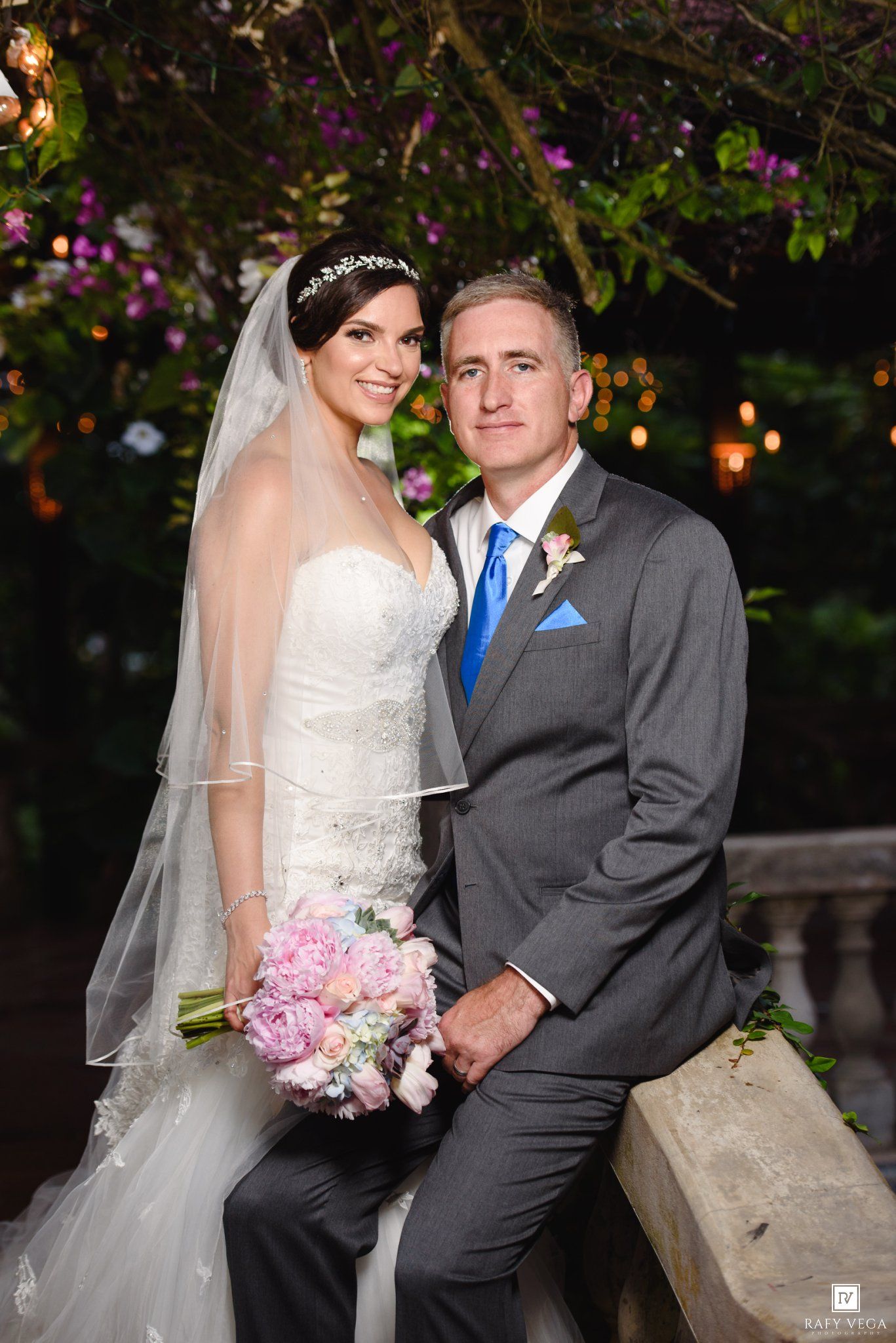 Bride in white gown holding pink bouquet poses with groom in gray suit and blue tie at outdoor wedding.