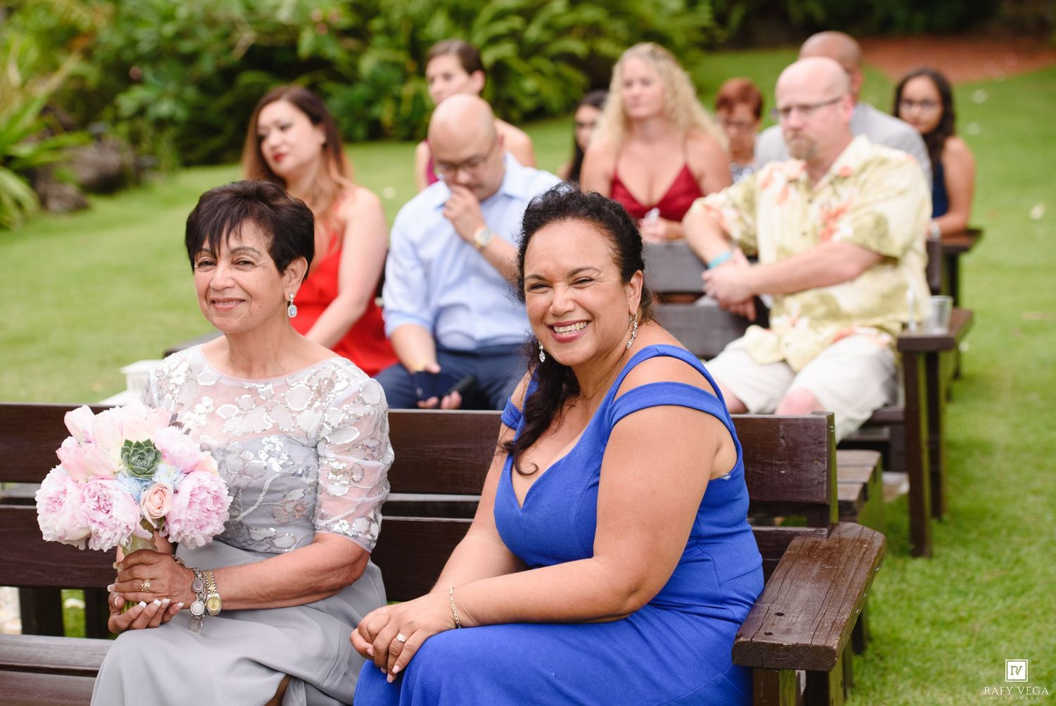 Two smiling women sit on wooden benches as guests at an outdoor garden wedding ceremony.
