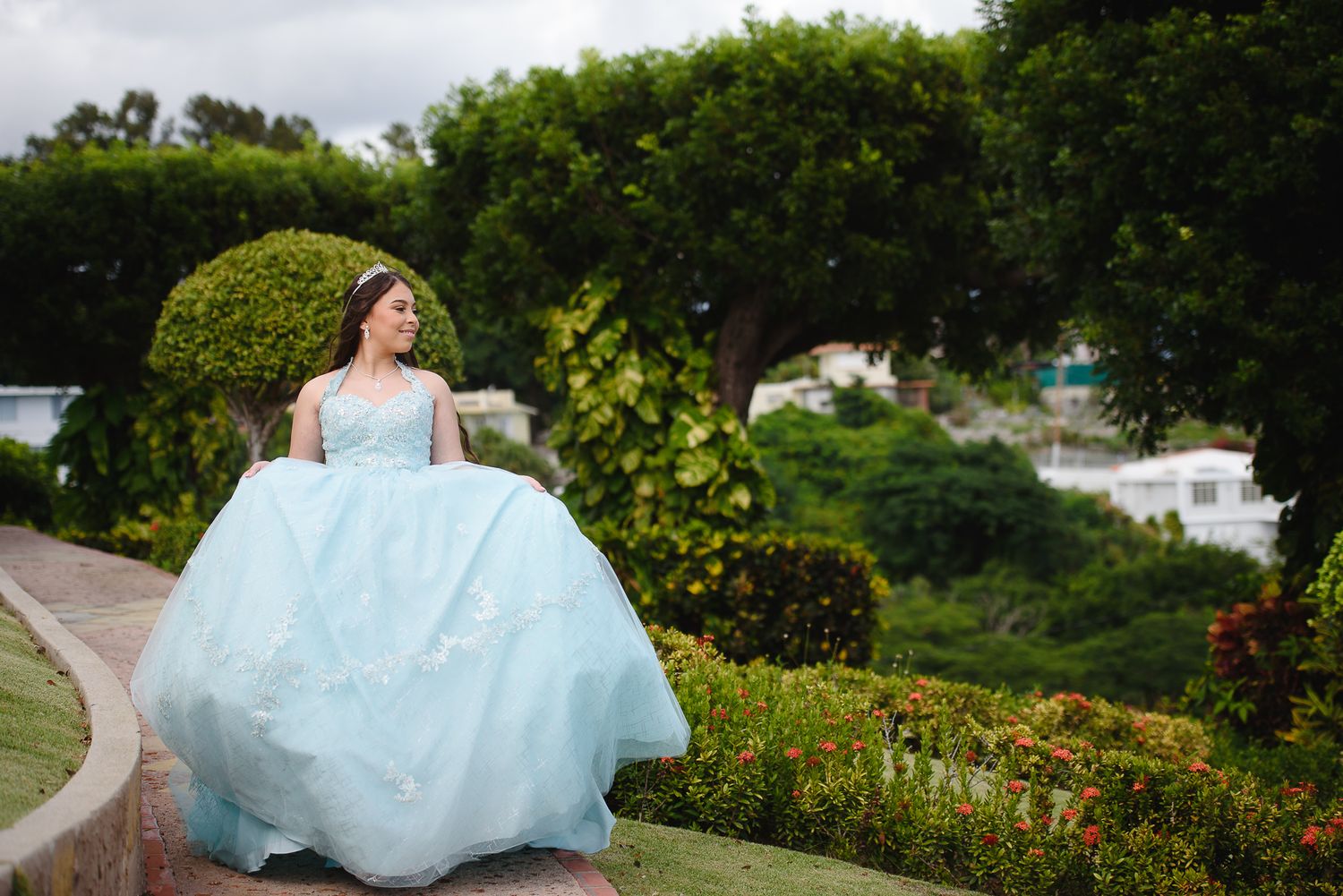 Young woman in a light blue ball gown poses gracefully in a lush garden setting for a quinceañera celebration.
