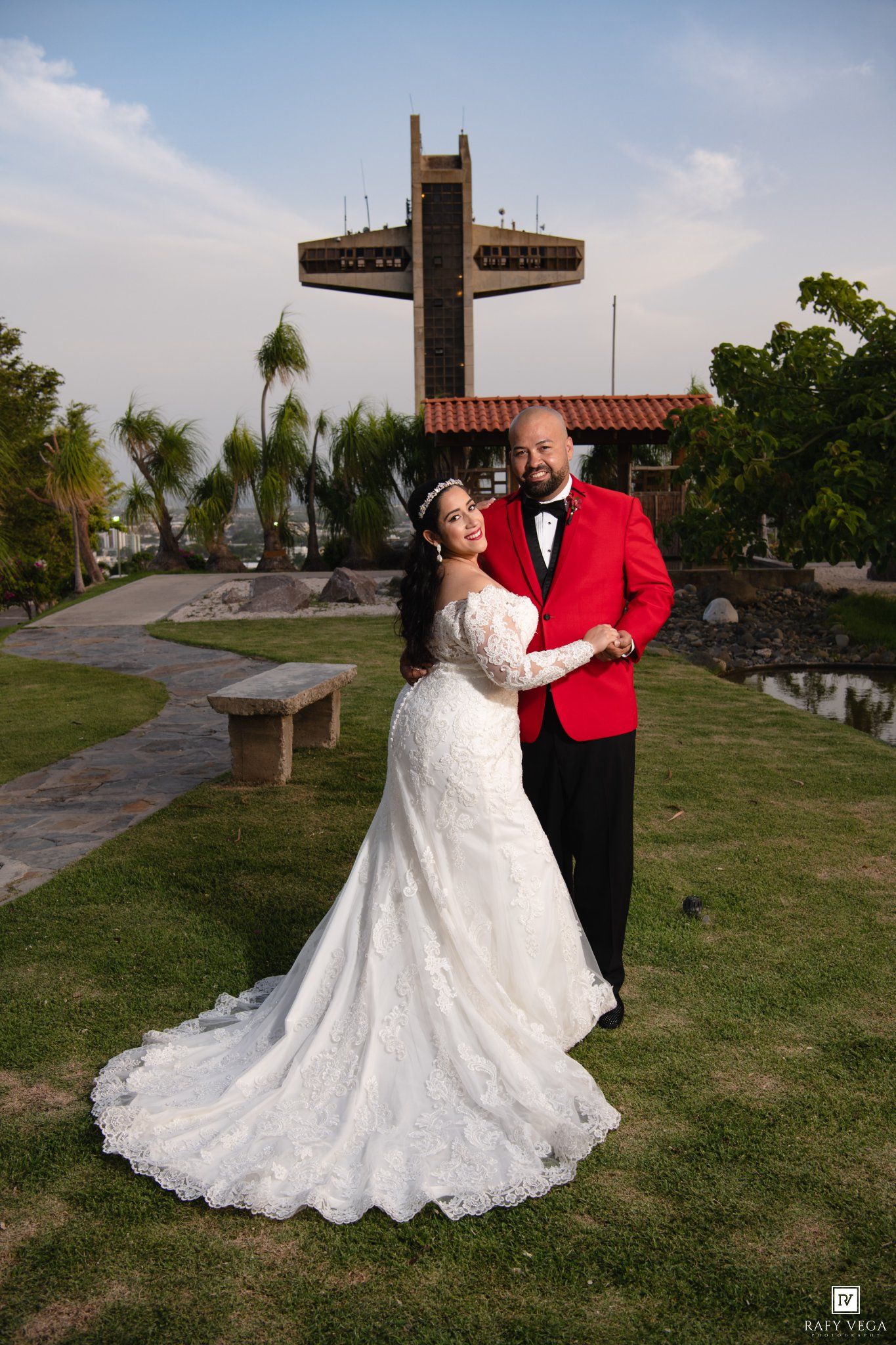 Bride in white lace gown and groom in red tuxedo pose together on green lawn with large cross monument behind them.