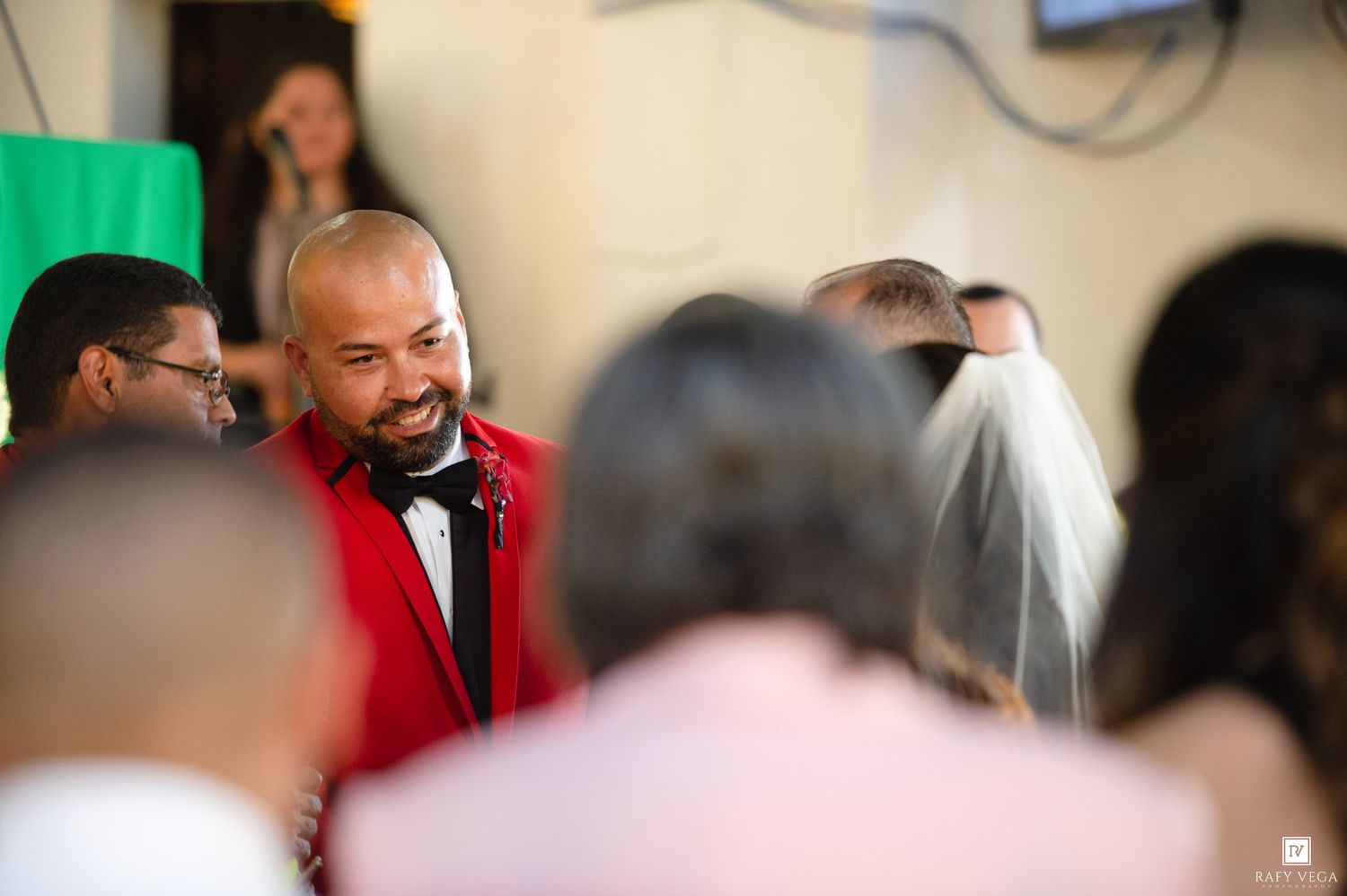 A smiling groom in a red suit and black bow tie mingles with wedding guests, with a bride in a veil visible nearby.