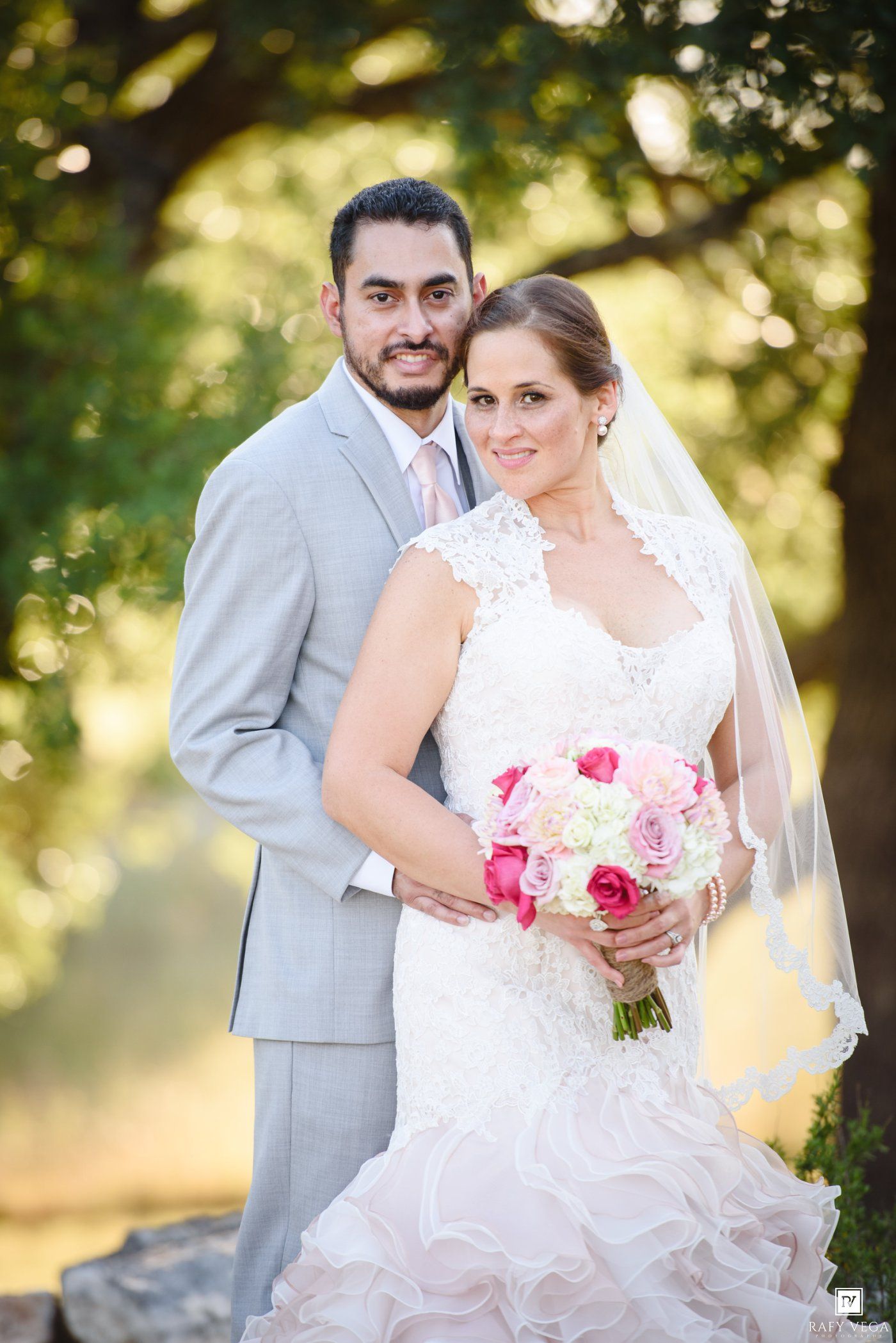 Bride in lace gown holding pink bouquet poses with groom in gray suit outdoors under trees on wedding day.