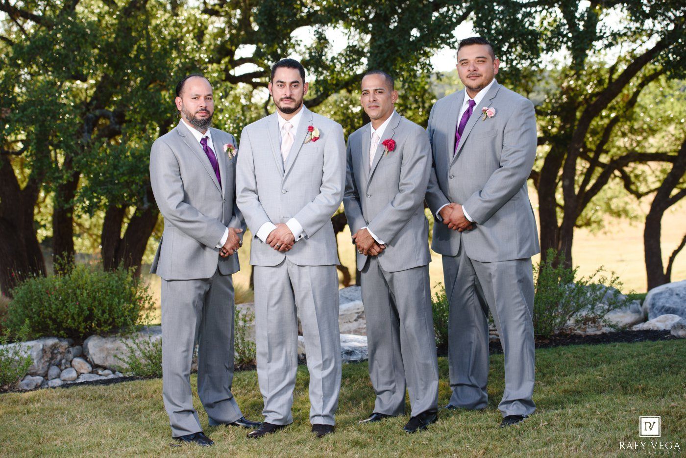 Four groomsmen in gray suits with purple boutonnieres posing outdoors at a wedding ceremony.