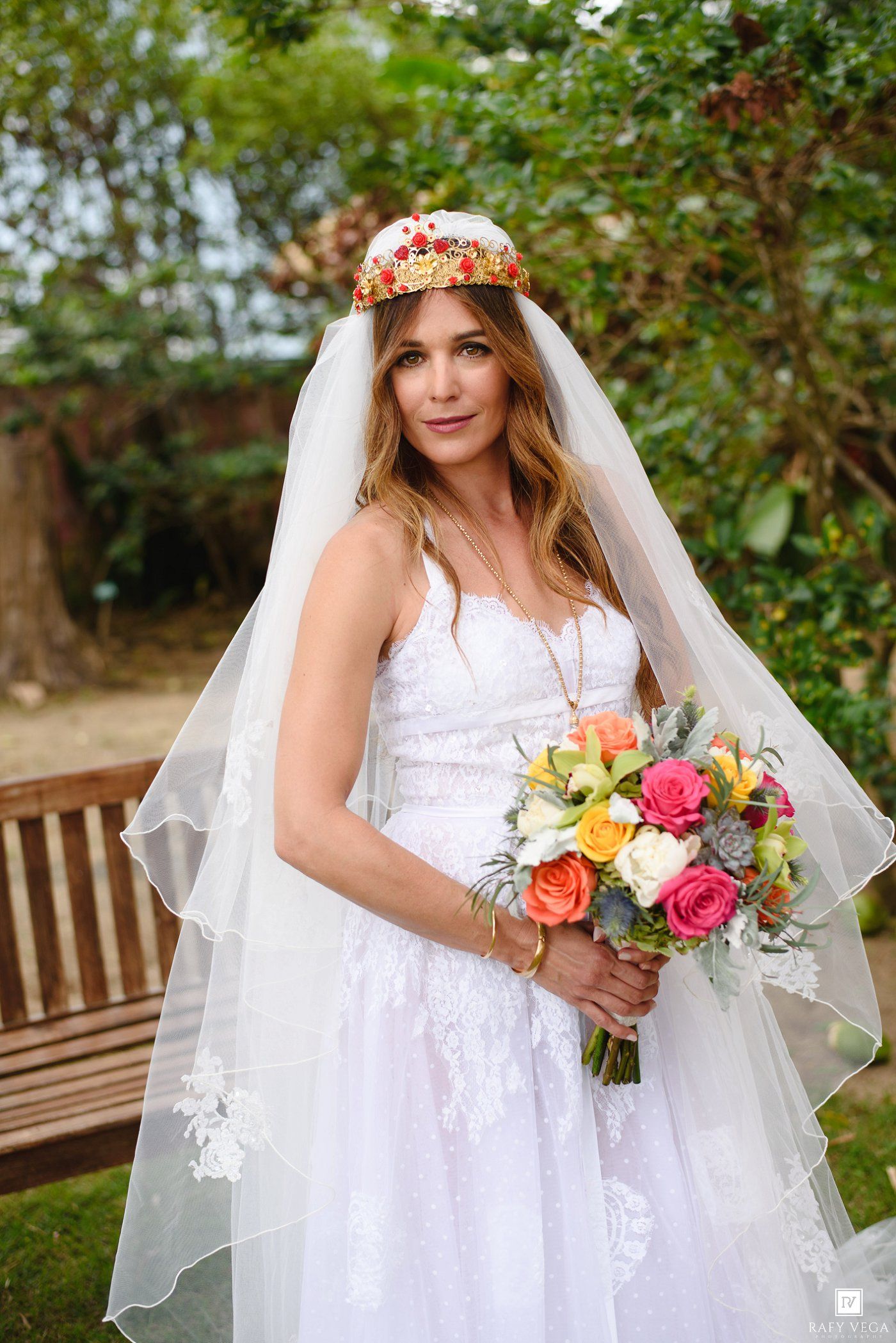 Bride in white lace gown and floral crown holds colorful bouquet of roses outdoors.