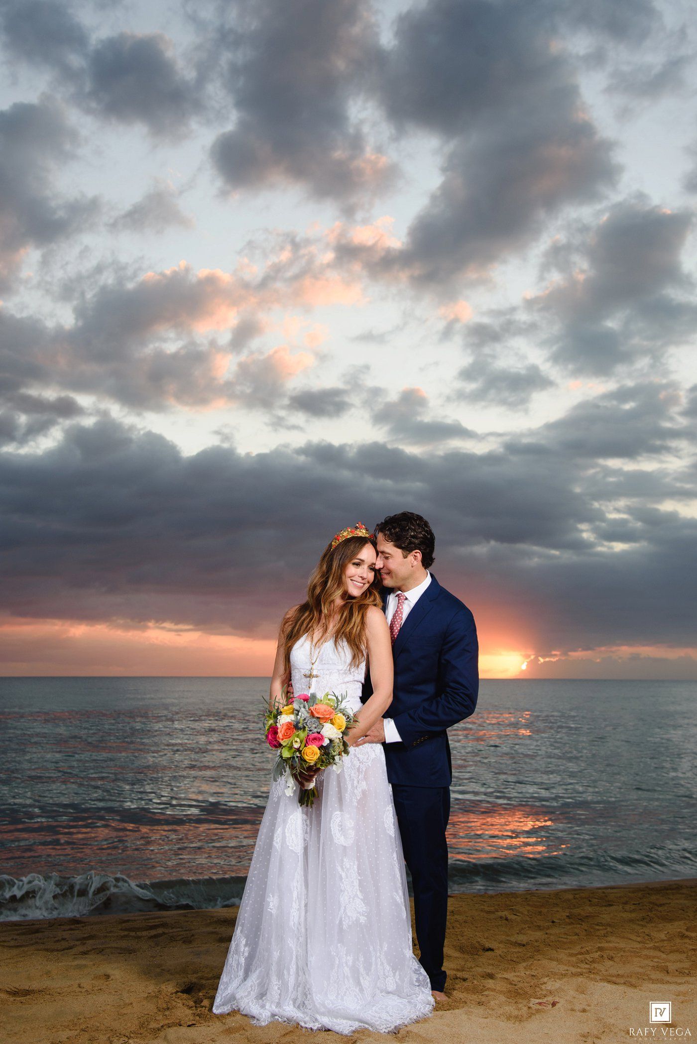 Bride and groom embrace on beach at sunset, she holds colorful bouquet, dramatic cloudy sky over ocean backdrop.